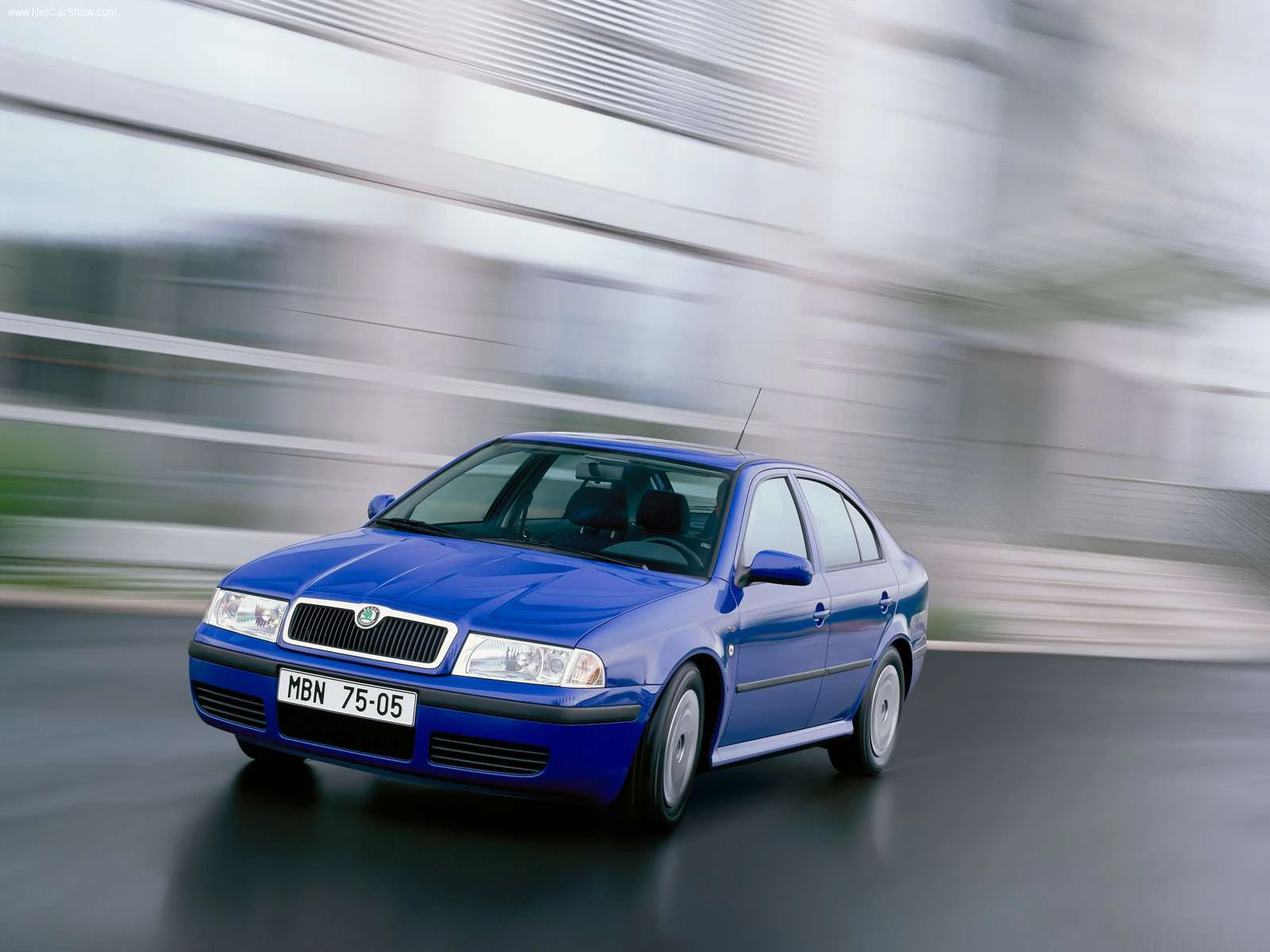 Blauer Skoda Octavia Limousine fährt mit Geschwindigkeit auf einer nassen Straße, modernes Gebäude im Hintergrund.