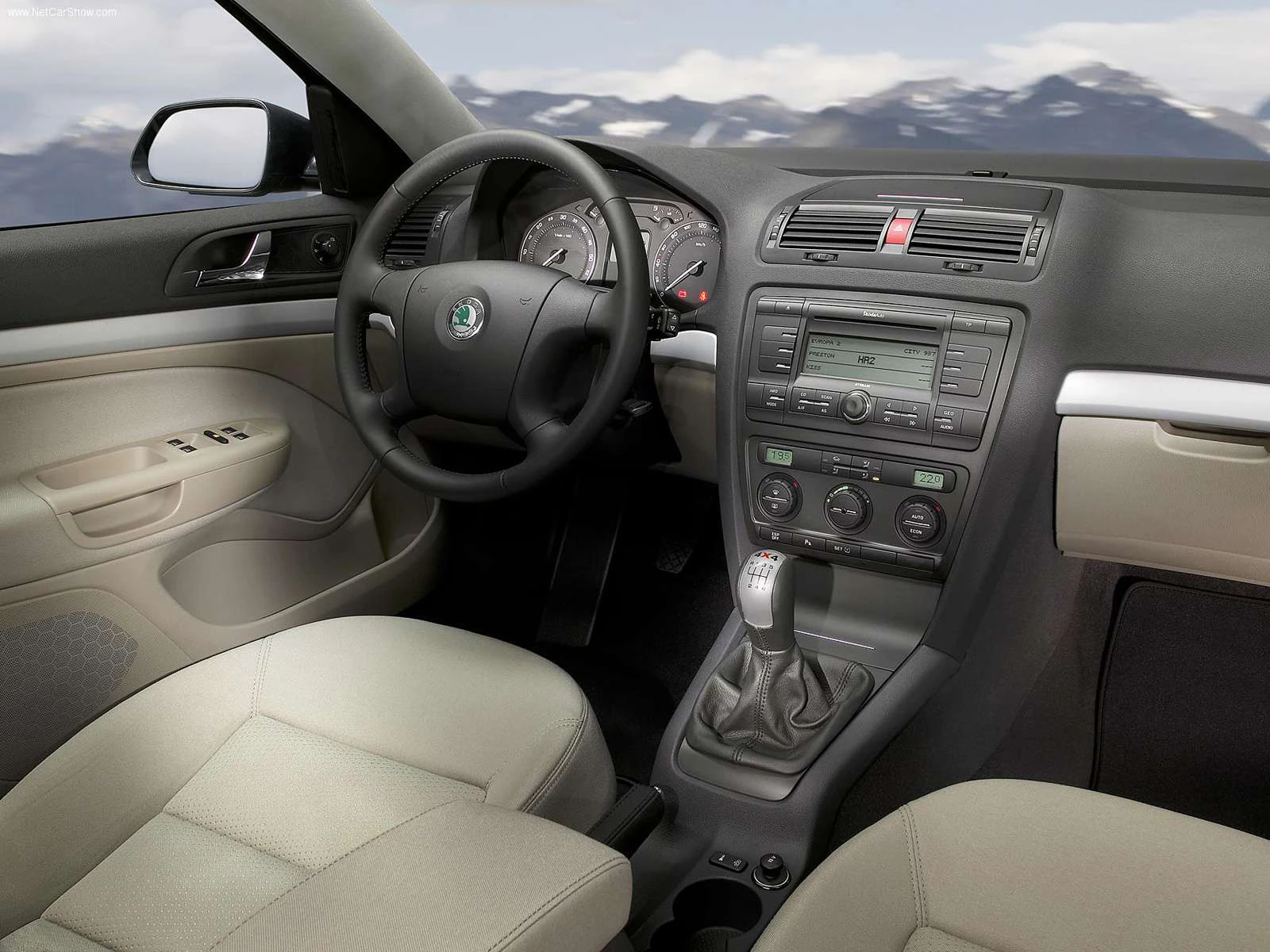 Innenraum eines Autos mit beigen Sitzen, Lenkrad, Armaturenbrett und Schaltknüppel, vor einer Berglandschaft im Hintergrund.