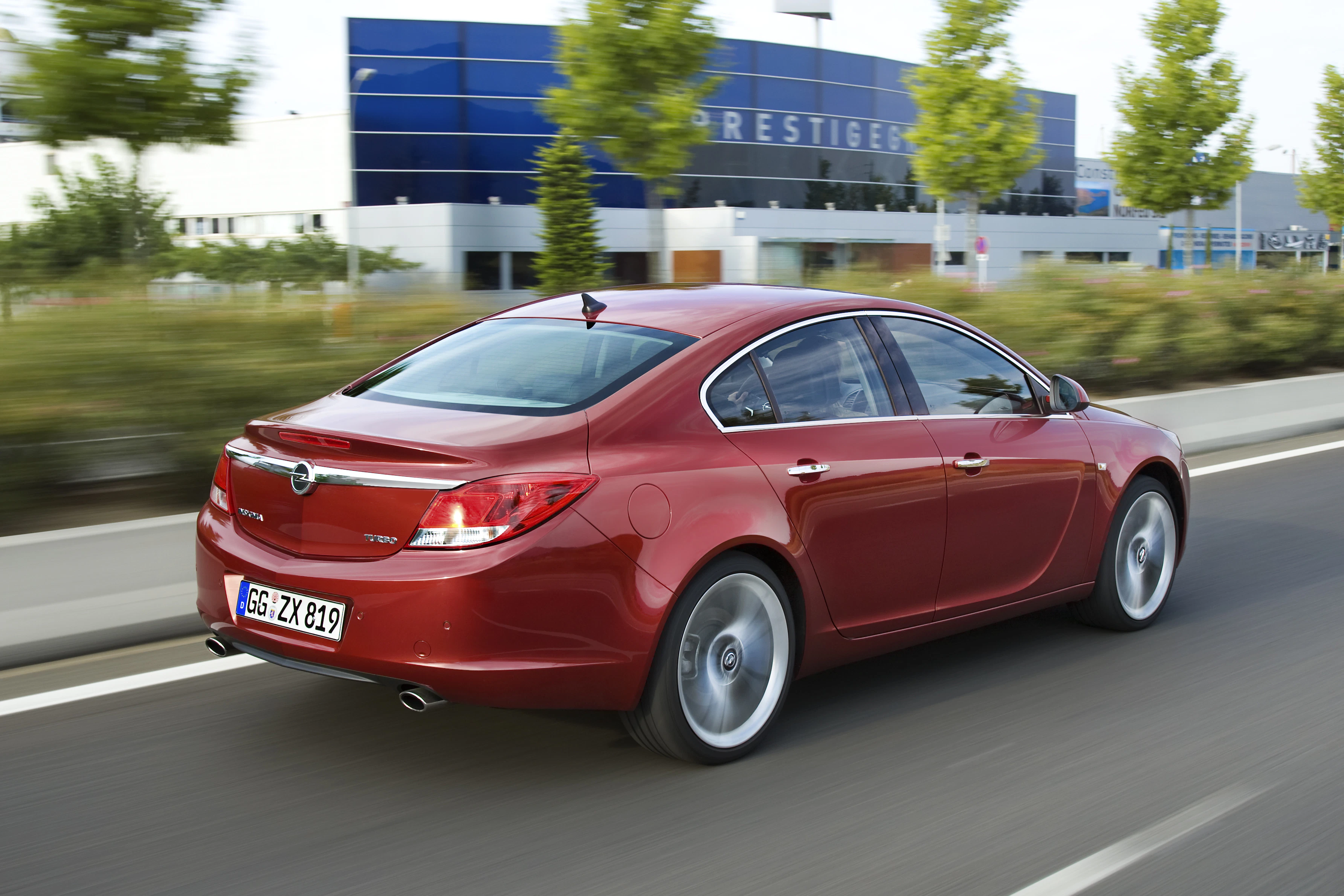 Roter Opel Insignia fährt auf einer Straße. Im Hintergrund sind Bäume und ein blaues Gebäude zu sehen.