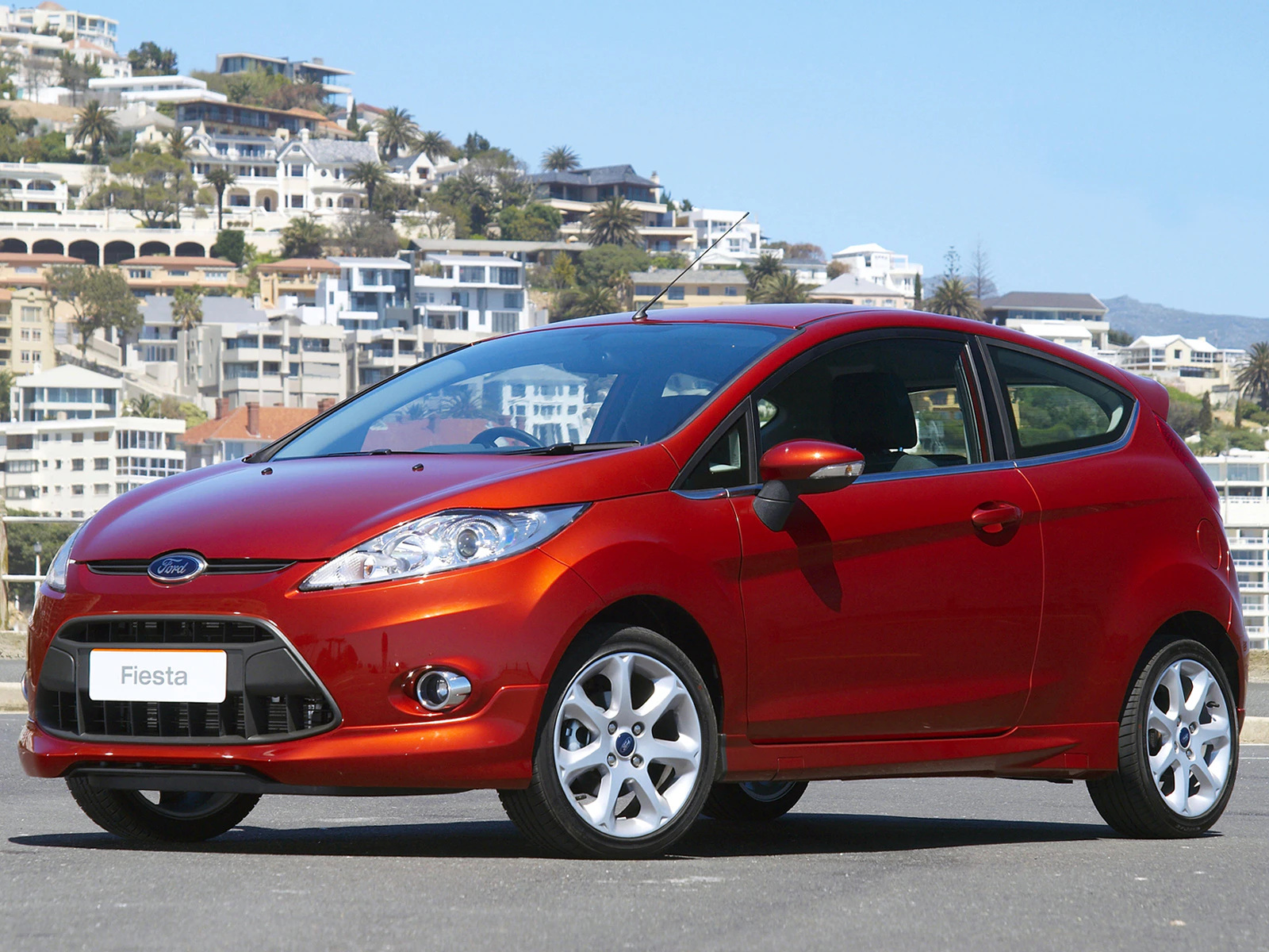Roter Ford Fiesta geparkt vor einer malerischen Küstenstadt mit weißem Wohnhäusern im Hintergrund an einem sonnigen Tag.