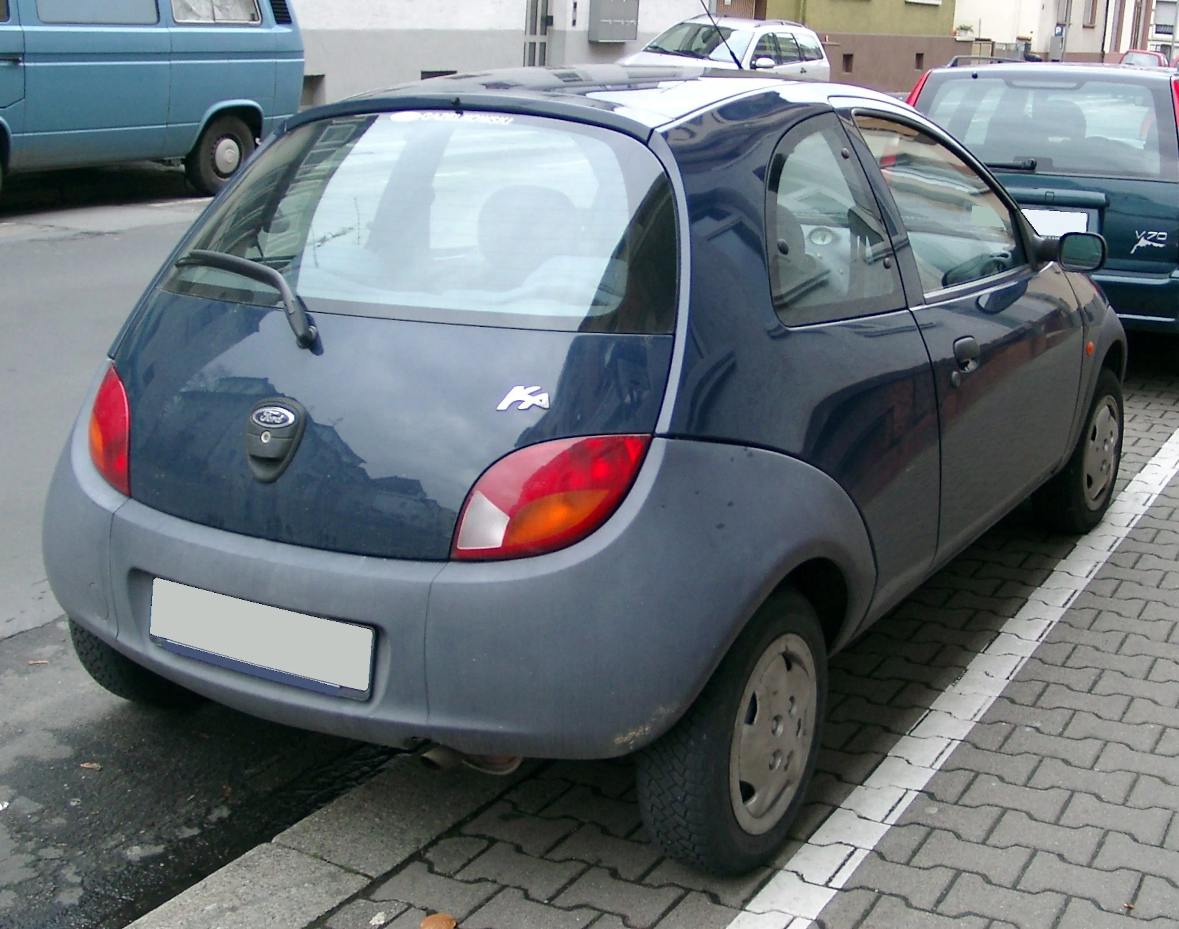 Blauer Ford Ka parkt rückwärts auf einer Straße, leicht schräg zur Bordsteinkante, mit umliegenden Fahrzeugen im Hintergrund.