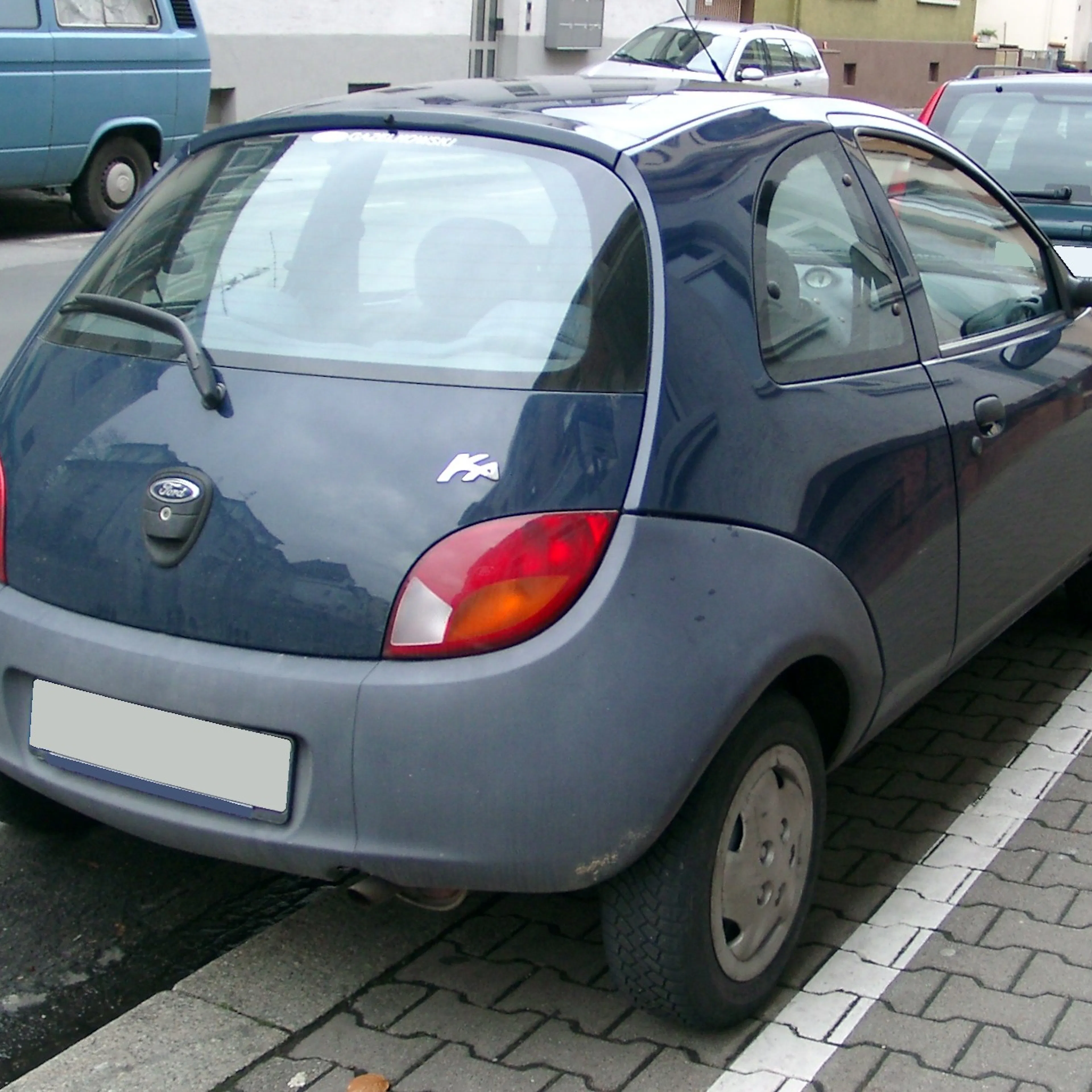 Blauer Ford Ka parkt rückwärts auf einer Straße, leicht schräg zur Bordsteinkante, mit umliegenden Fahrzeugen im Hintergrund.
