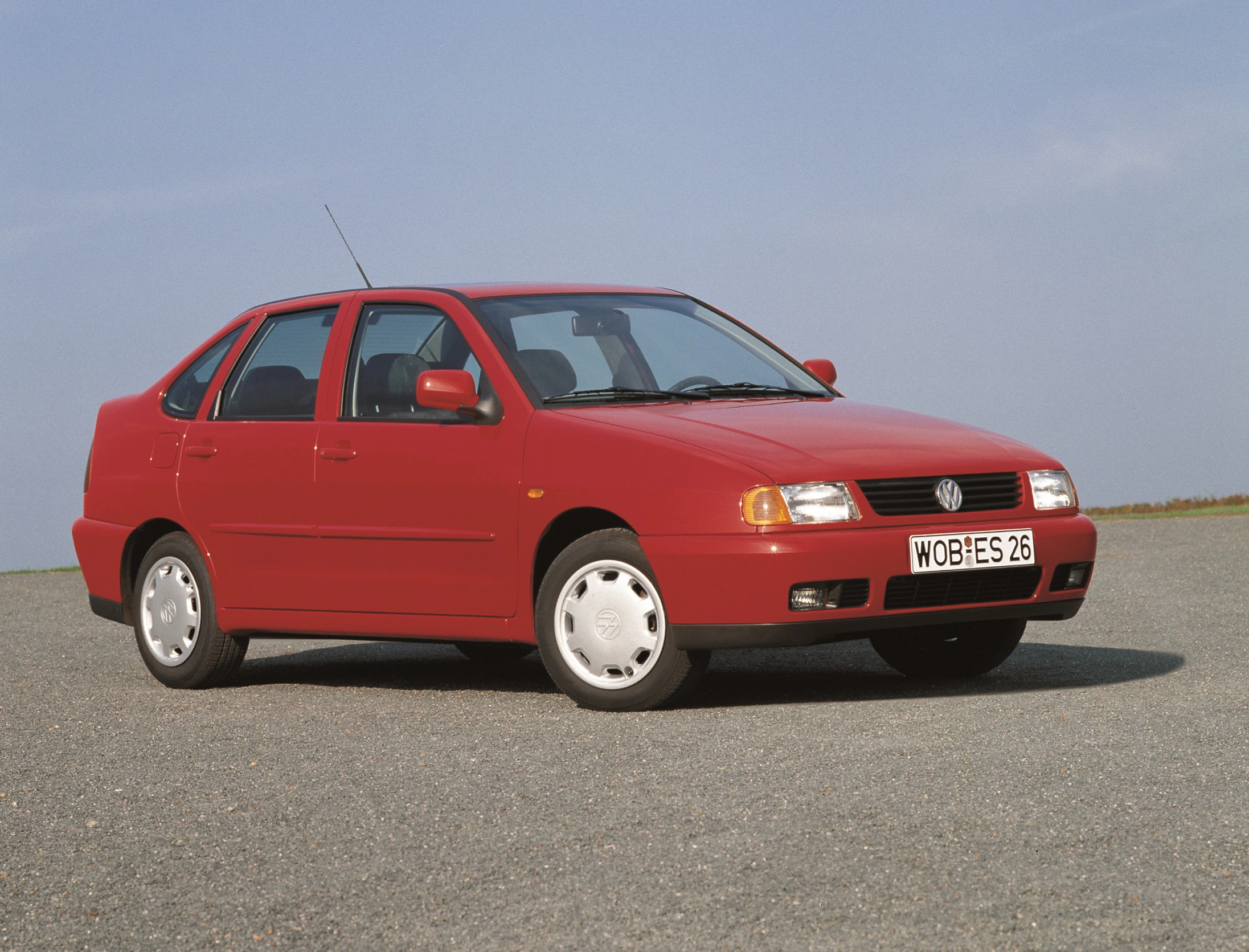 Roter VW Polo Classic der 1990er Jahre, seitlich von vorne fotografiert, auf einem asphaltierten Platz unter klarem Himmel.