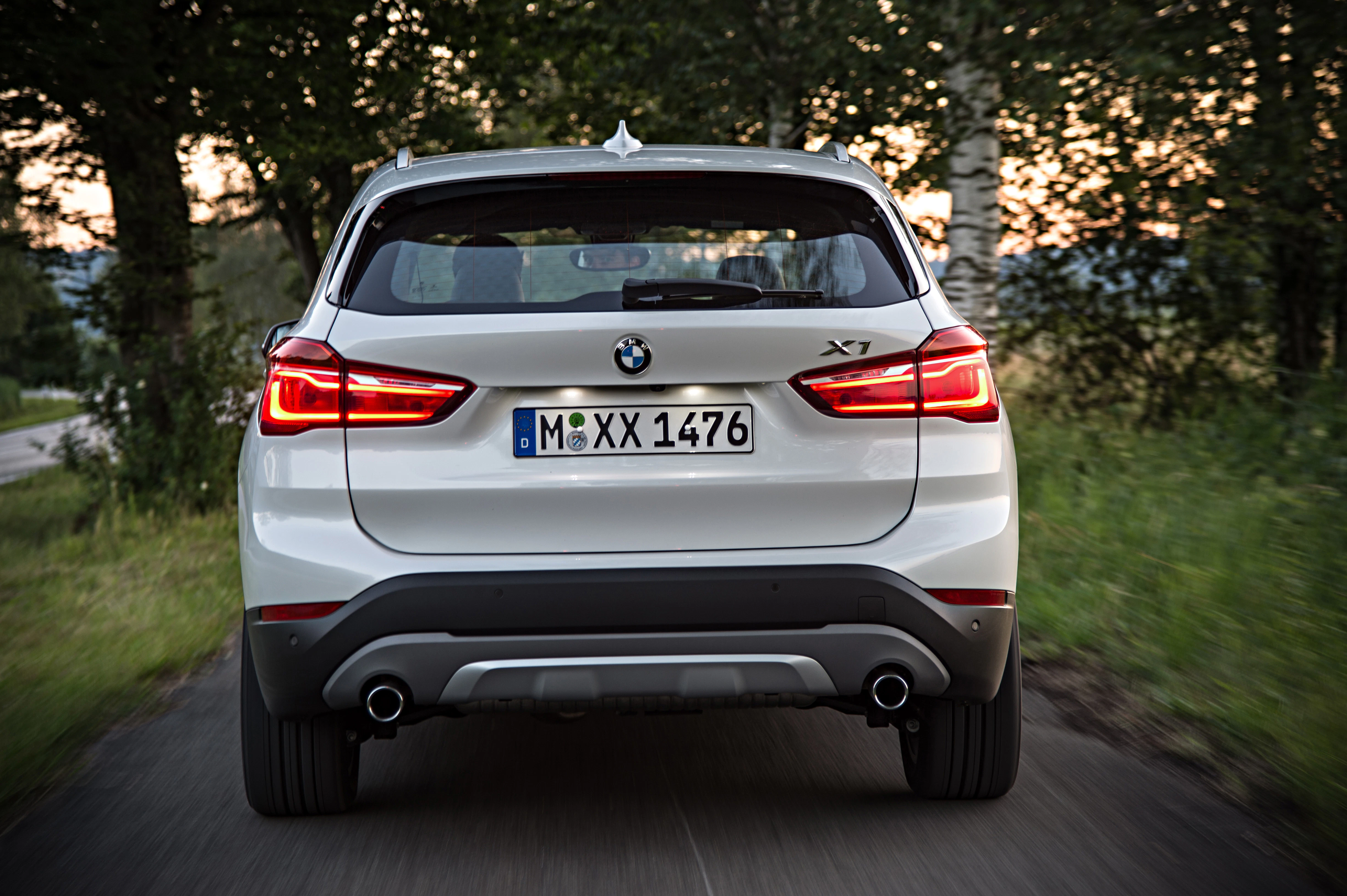 Rückansicht eines weißen BMW X1 auf einer Landstraße bei Sonnenuntergang, umgeben von Bäumen.