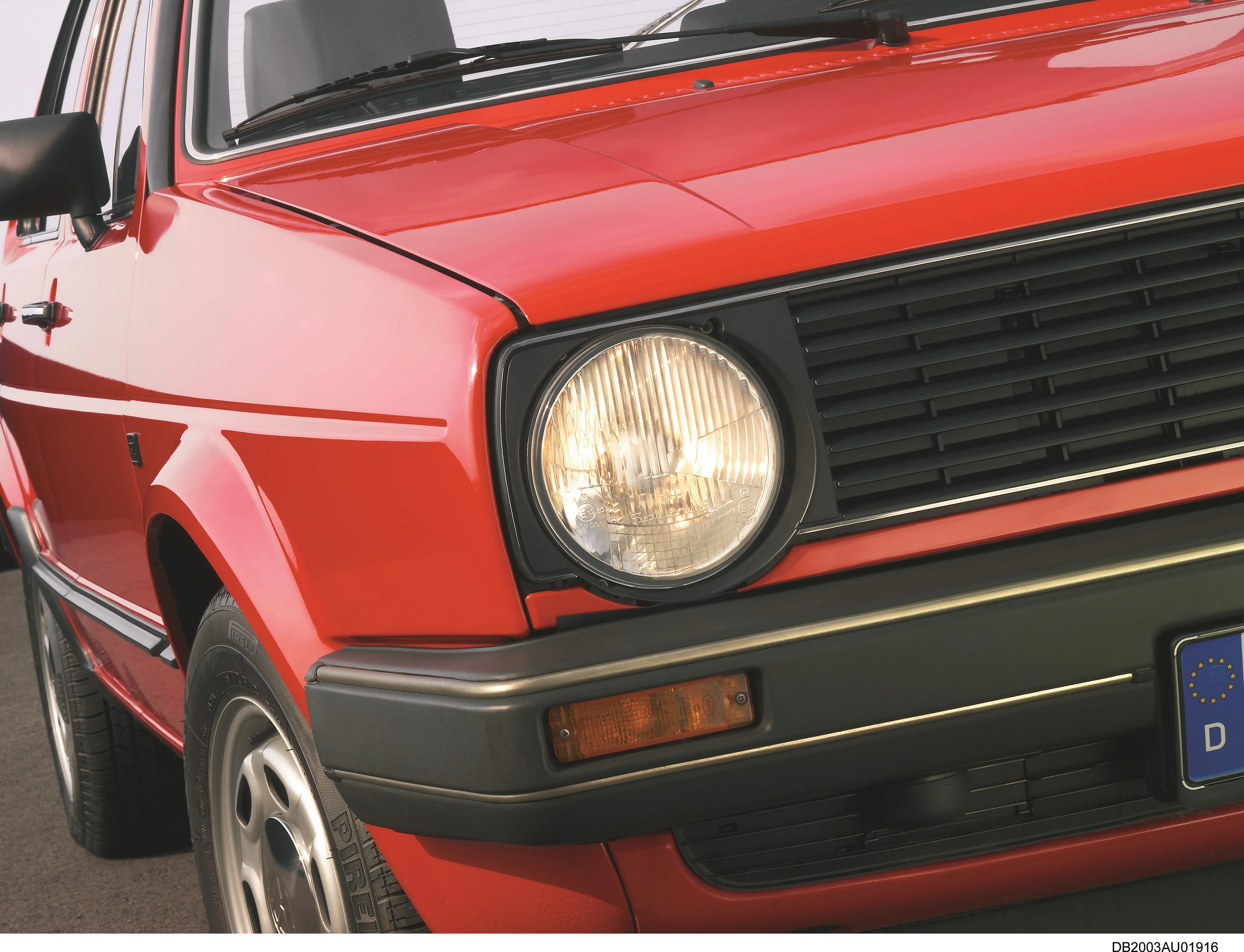 Roter, klassischer Kleinwagen von vorne fotografiert, im Vordergrund der linke Frontscheinwerfer und ein Teil des Stoßfängers.