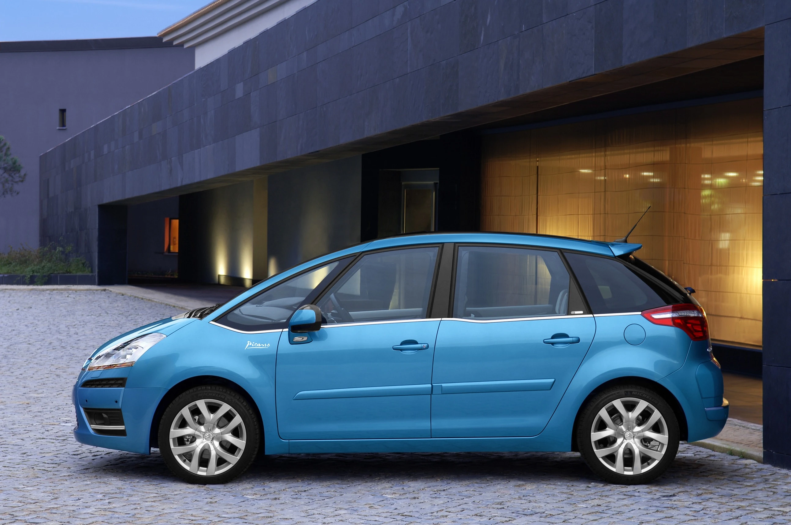 Ein blauer Citroën C4 Picasso steht auf einem gepflasterten Hof vor einem modernen Gebäude mit großen Fenstern.