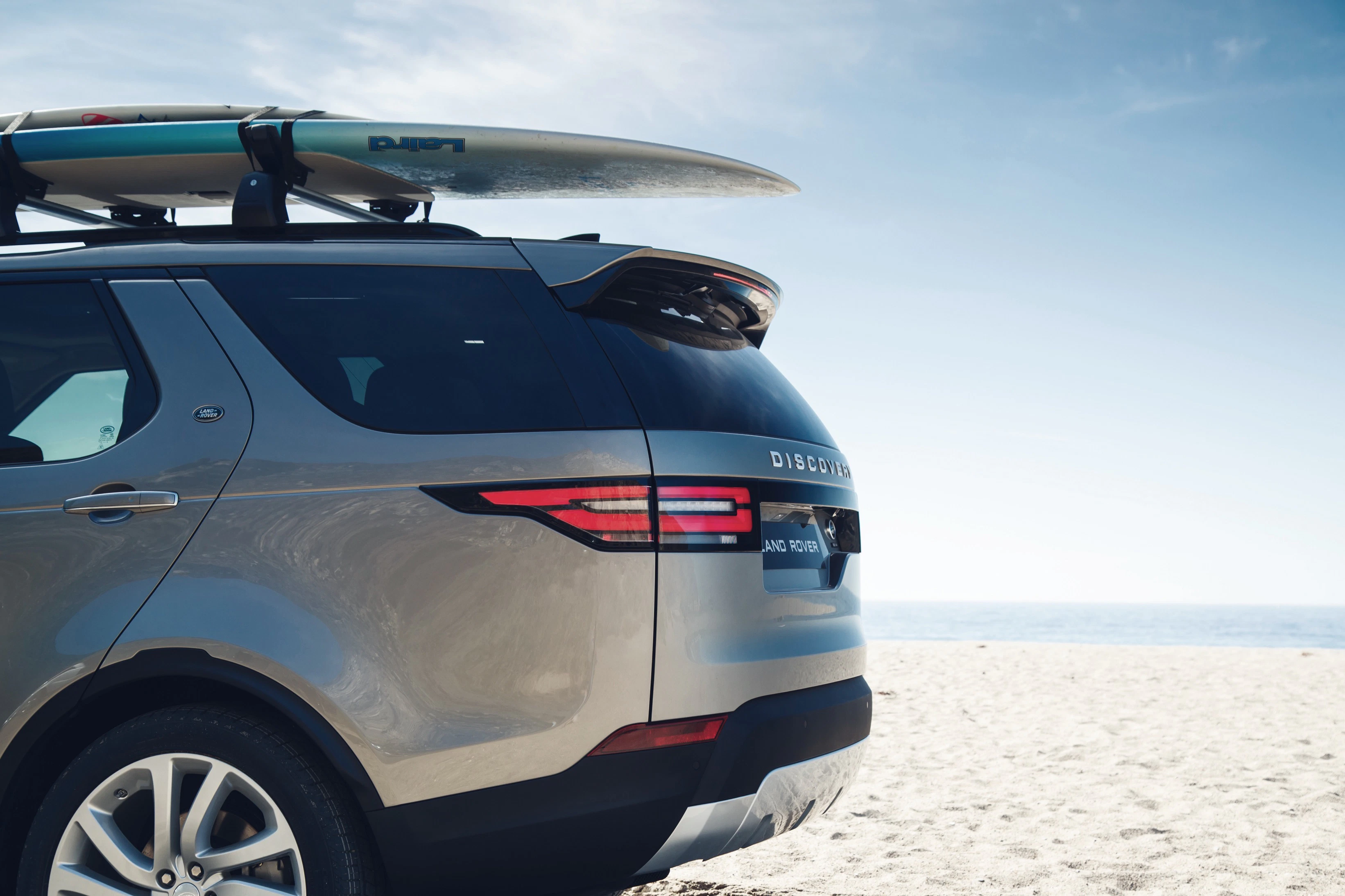 SUV mit Surfbrett auf dem Dach steht am Sandstrand. Der Himmel ist klar und blau, im Hintergrund das Meer.