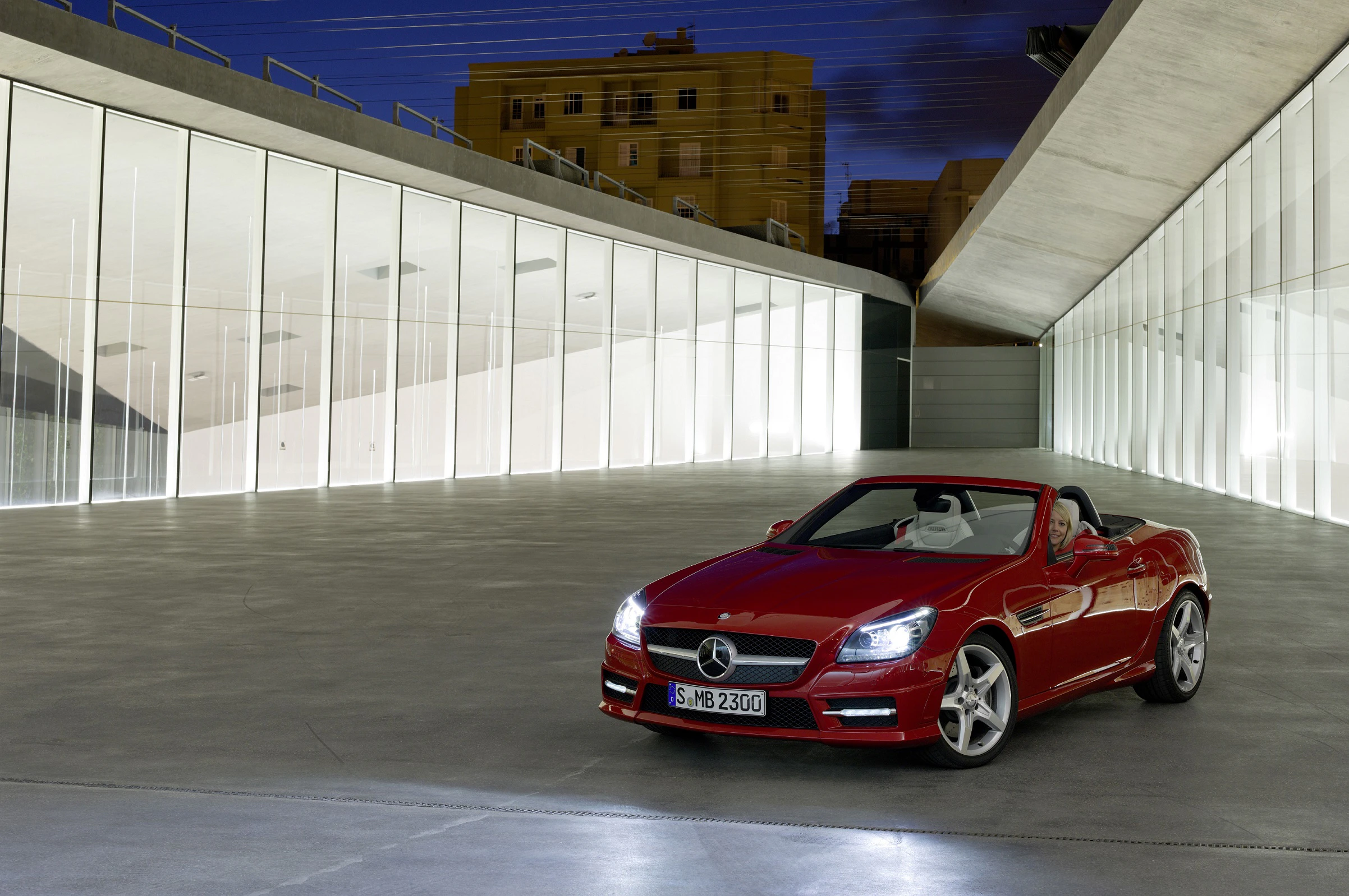 Roter Mercedes-Benz SLK steht nachts in modern beleuchtetem, gläsernem Gebäude mit sternklarem Himmel im Hintergrund.