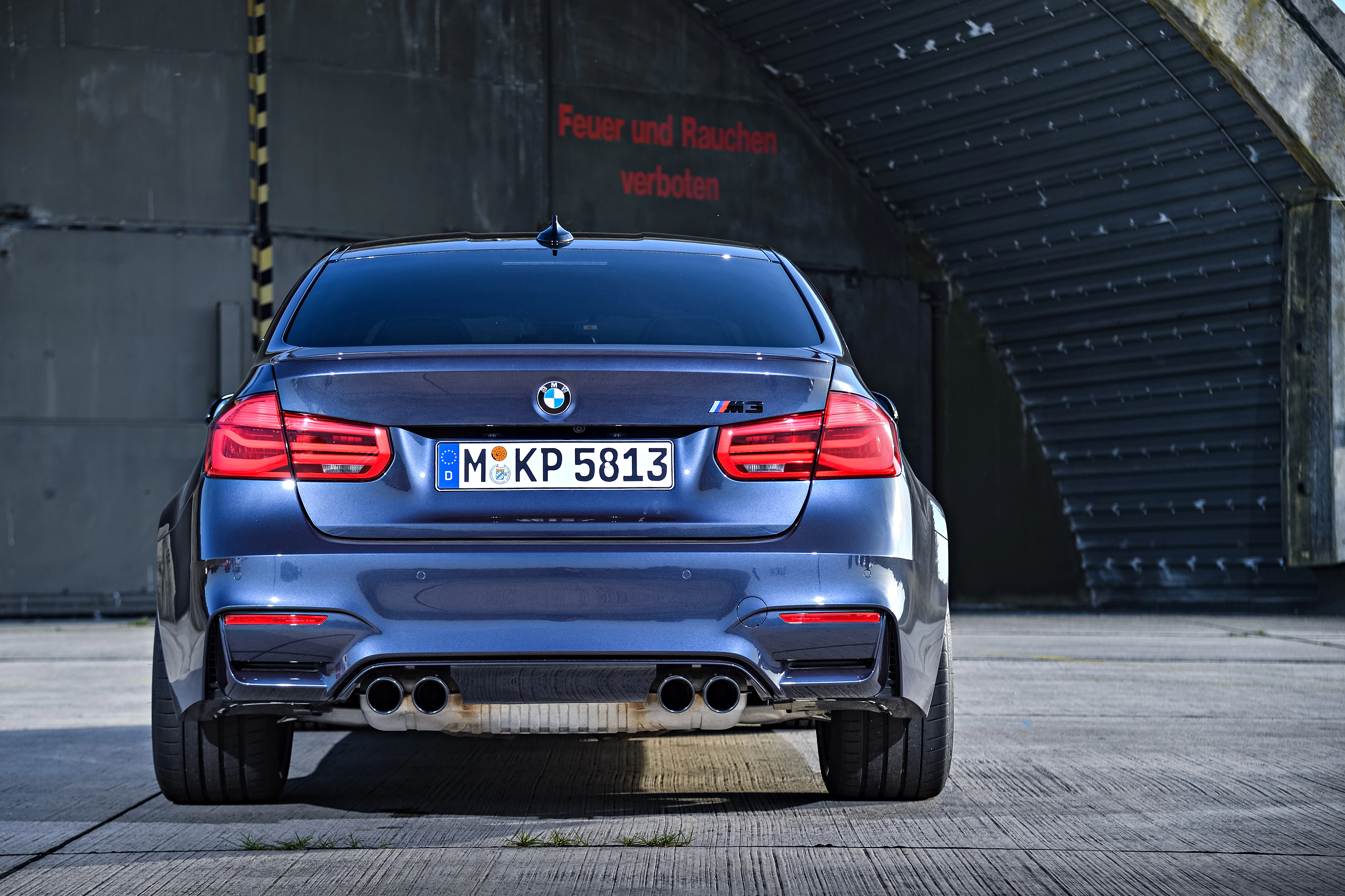Rückansicht eines grauen BMW M3 vor einem Hangar. Kennzeichen sichtbar, sportliches Design, vier Auspuffrohre. Schild: "Feuer und Rauchen verboten".
