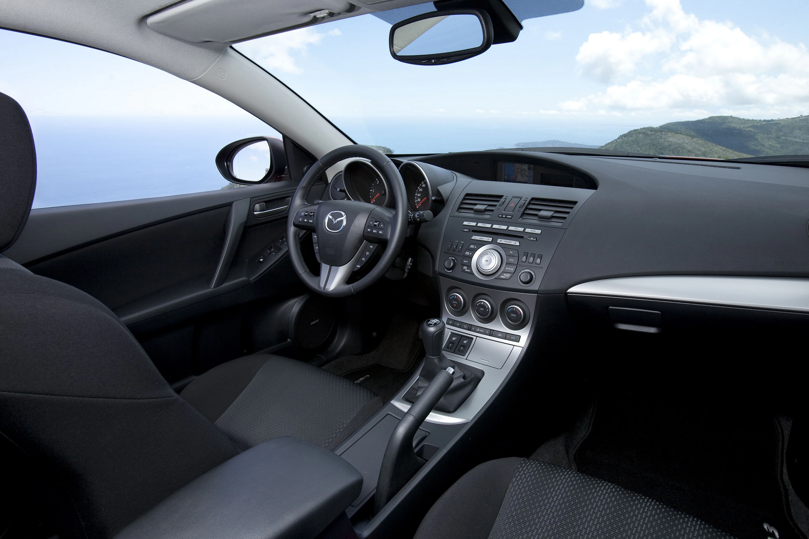 Innenraum eines Autos mit schwarzer Ausstattung, modernem Armaturenbrett, Lenkrad mit Logo, Schalthebel und Blick auf Meer und Himmel durch Fenster.