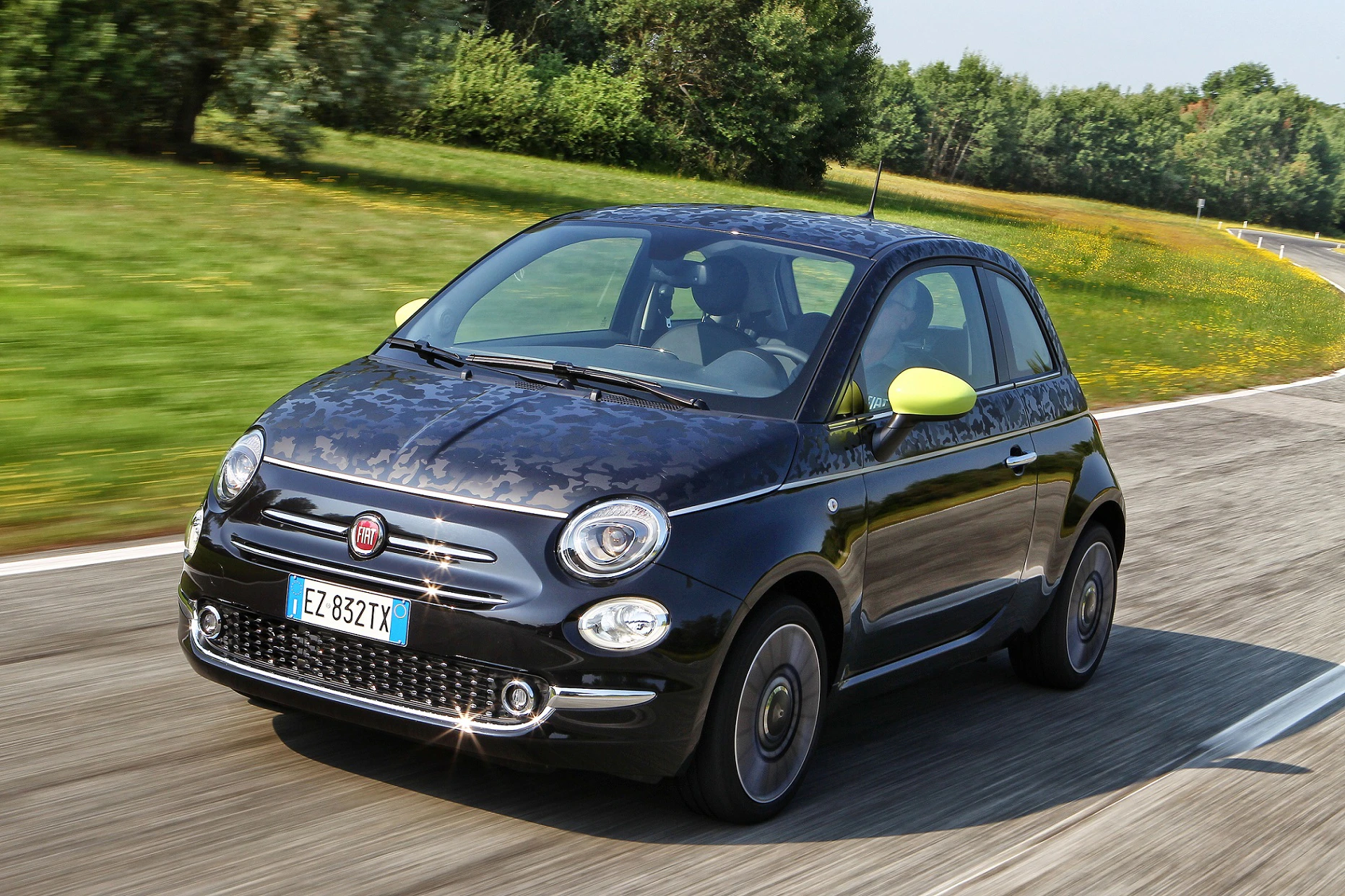 Kleiner schwarzer Fiat 500 mit gelben Seitenspiegeln fährt auf einer Landstraße. Im Hintergrund sind Blumenwiese und Bäume zu sehen.