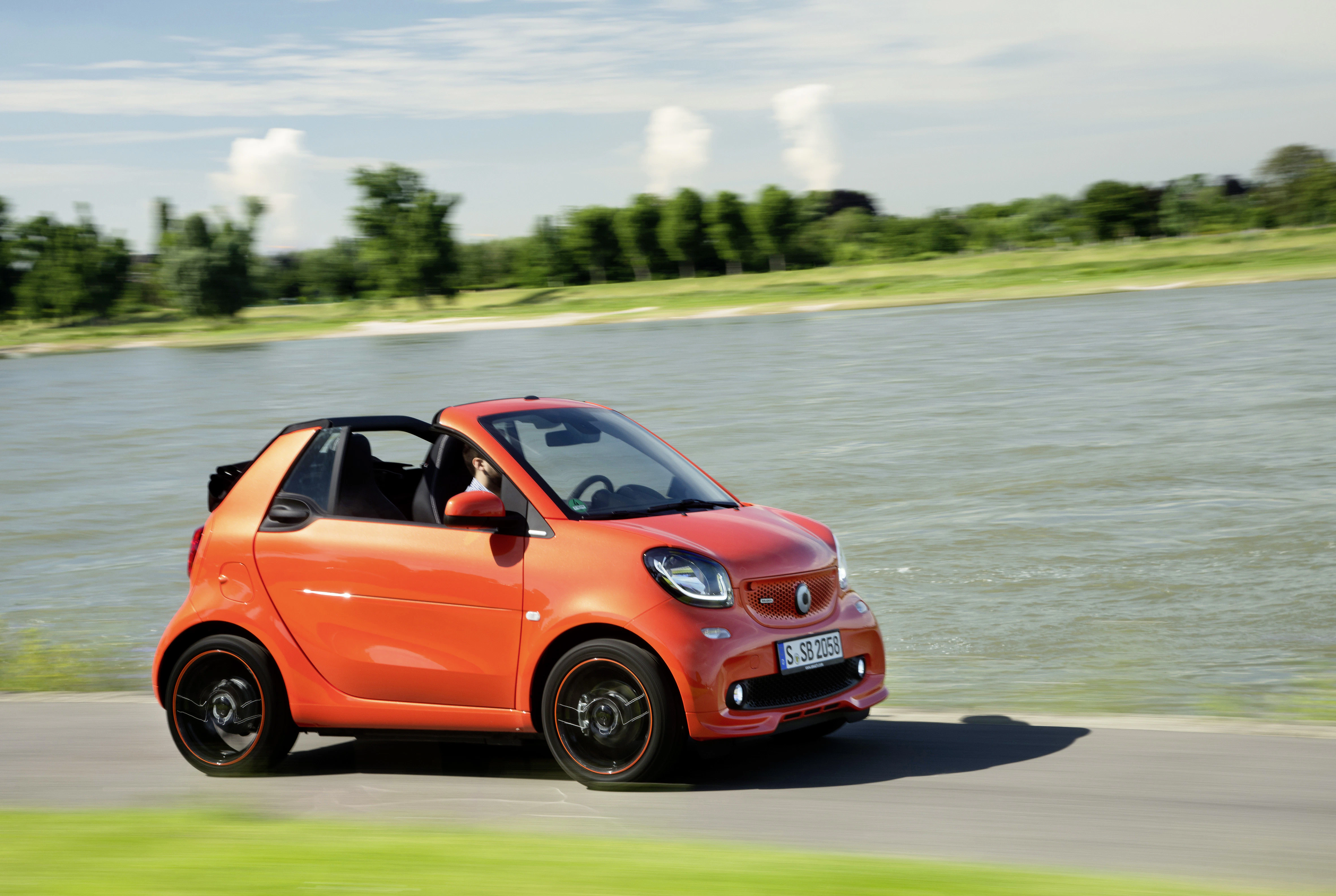 Roter Smart-Cabrio fährt auf einer Straße entlang eines Flusses, umgeben von grüner Landschaft und bewölktem Himmel im Hintergrund.