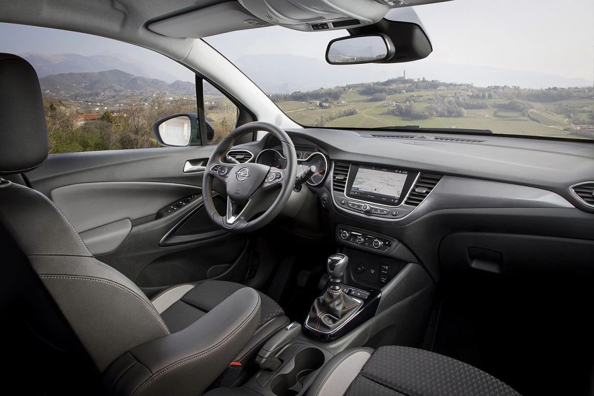 Innenraum eines Autos mit grauen Sitzen, modernem Armaturenbrett und Lenkrad. Durch die Windschutzscheibe sieht man eine ländliche Landschaft.