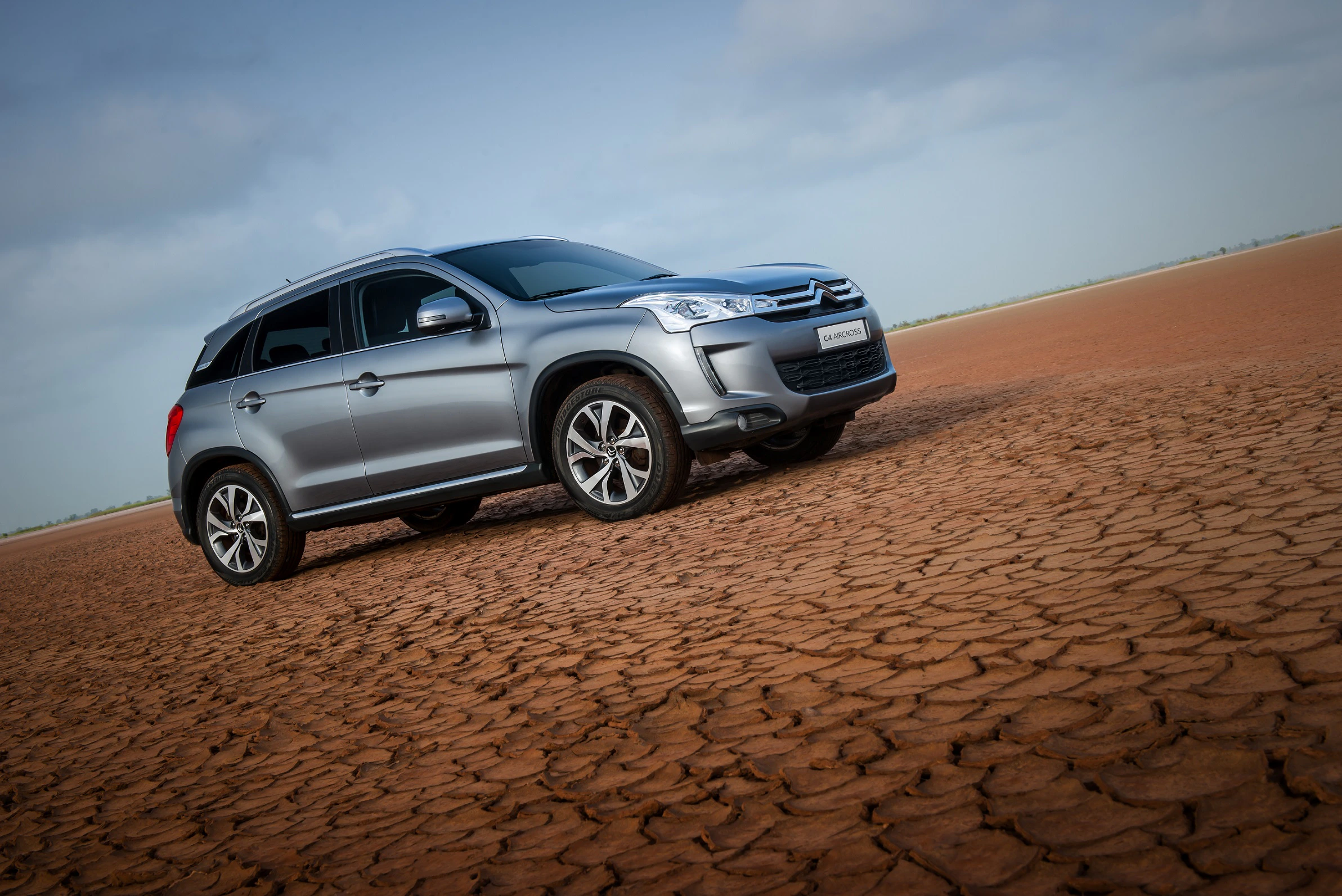 Silberner SUV parkt auf trockener, rissiger Erde unter blauem Himmel mit Wolken. Klare, helle Tageslichtaufnahme.
