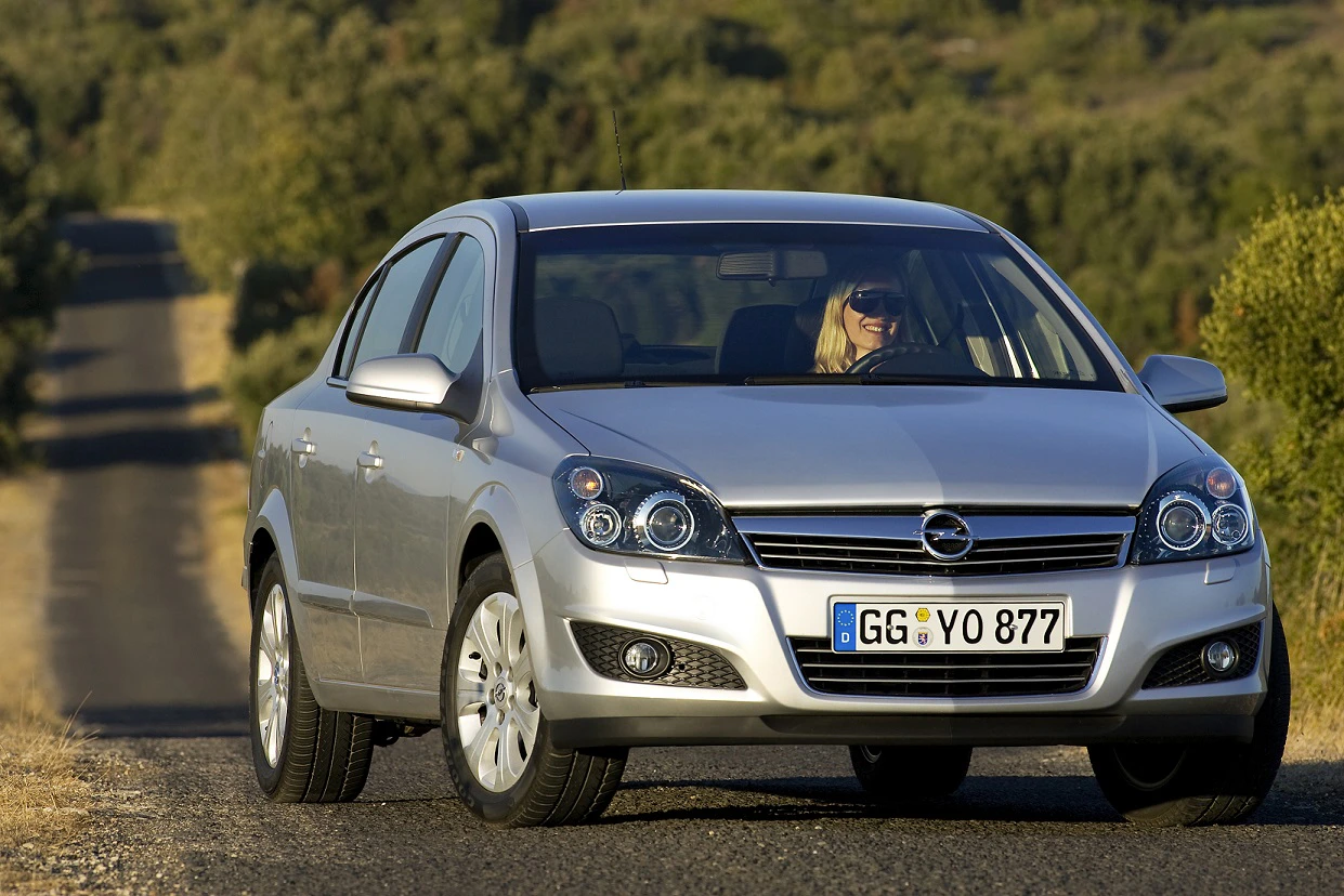 Silberner Opel Astra fährt auf einer ländlichen Straße, umgeben von Bäumen. Eine Person sitzt am Steuer bei sonnigem Wetter.