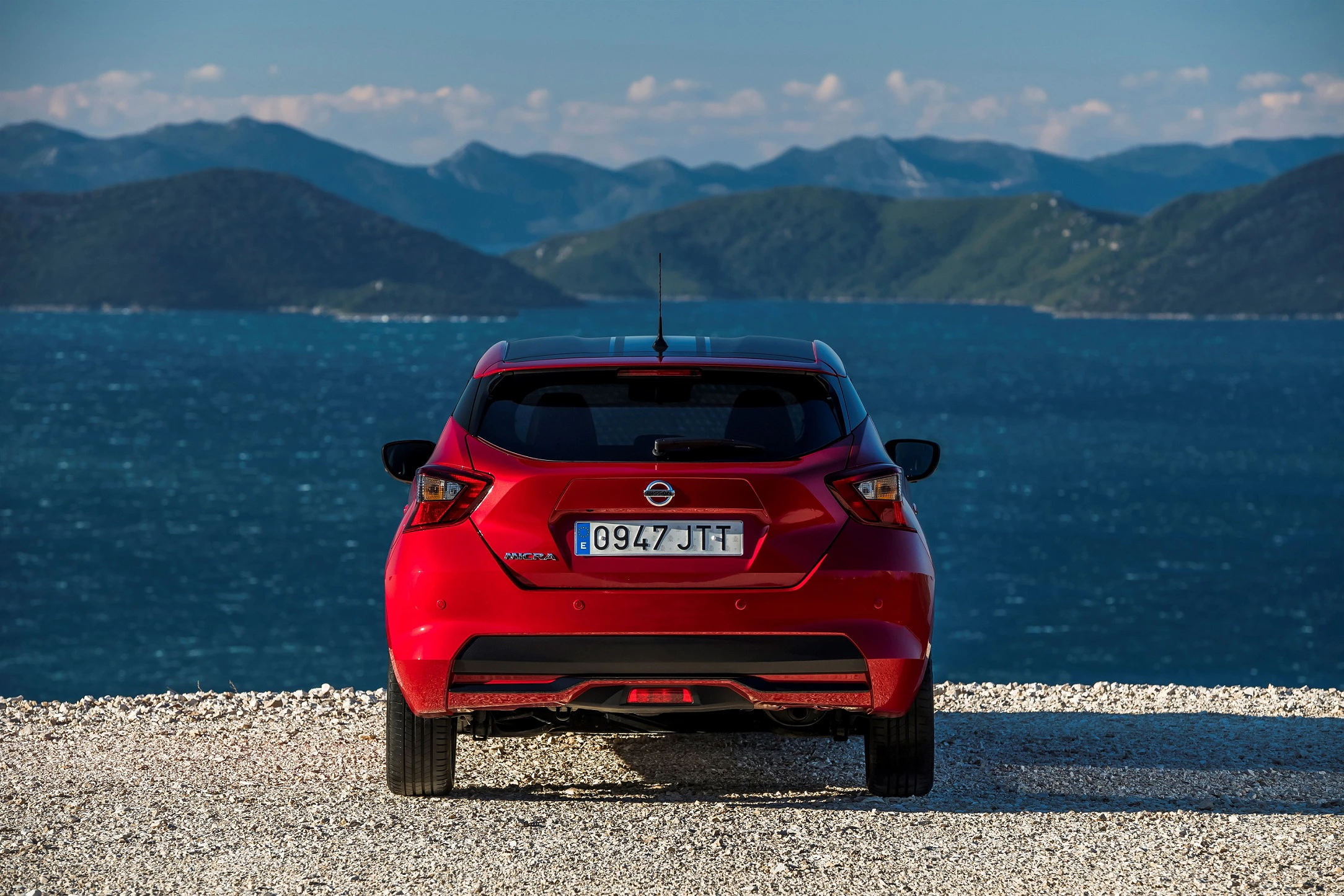 Rotes Auto parkt auf Kiesweg mit Blick auf ein blaues Meer und bergige Landschaft im Hintergrund.