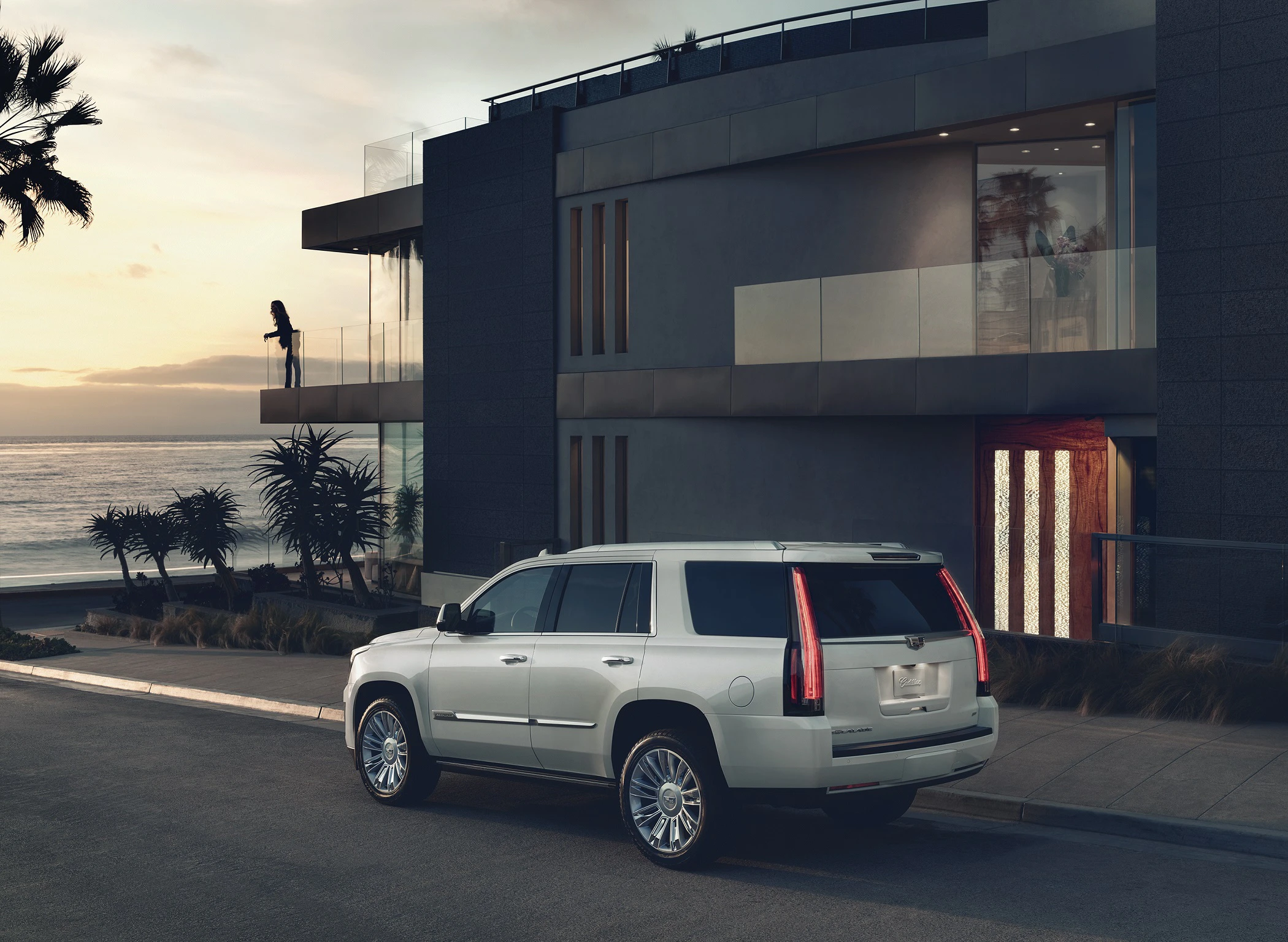 Weiße Luxus-SUV vor modernem Haus bei Sonnenuntergang, Palmen im Hintergrund, Blick auf das Meer.