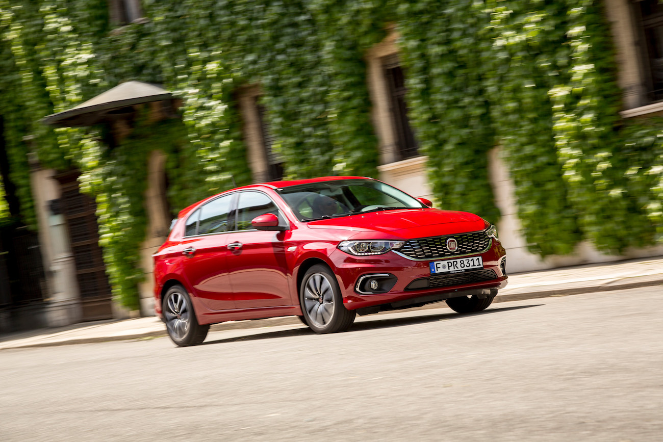 Roter Pkw fährt auf Straße vor einer mit Efeu bewachsenen Mauer entlang, im Hintergrund ein historisches Gebäude.