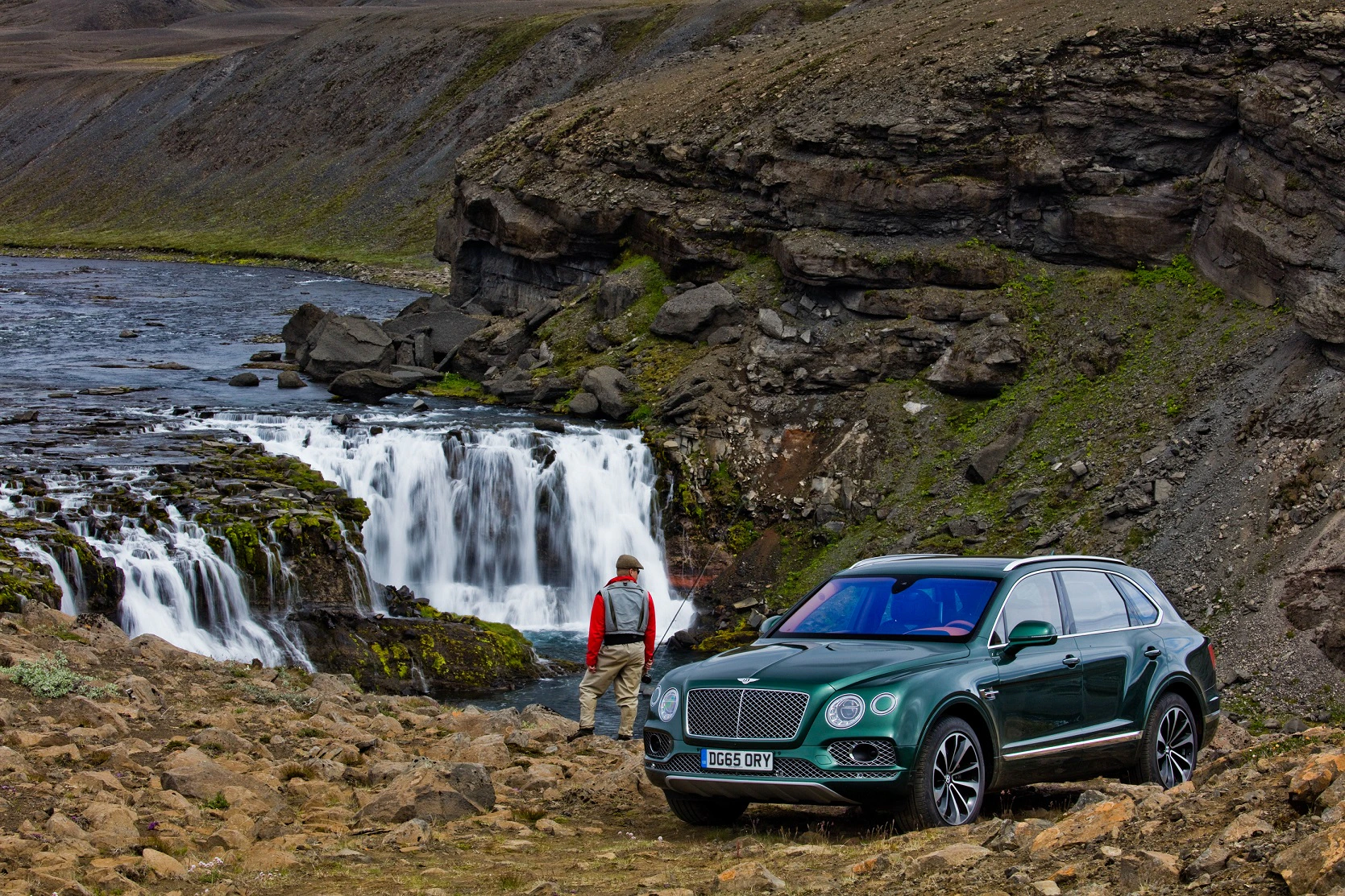 Ein grüner SUV parkt auf felsigem Gelände neben einem Wasserfall und einem Fluss, während eine Person in roter Jacke das Szenario betrachtet.