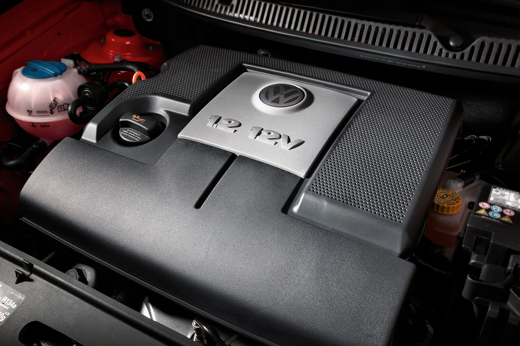 Nahaufnahme eines VW-Motors mit 12V-Kennzeichnung und schwarzer Abdeckung im Motorraum eines Fahrzeugs.