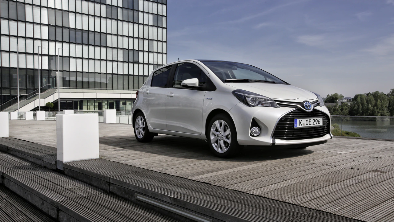 Silberner Toyota Yaris Hybrid parkt auf Holzplattform vor modernem Gebäude, blauer Himmel im Hintergrund.