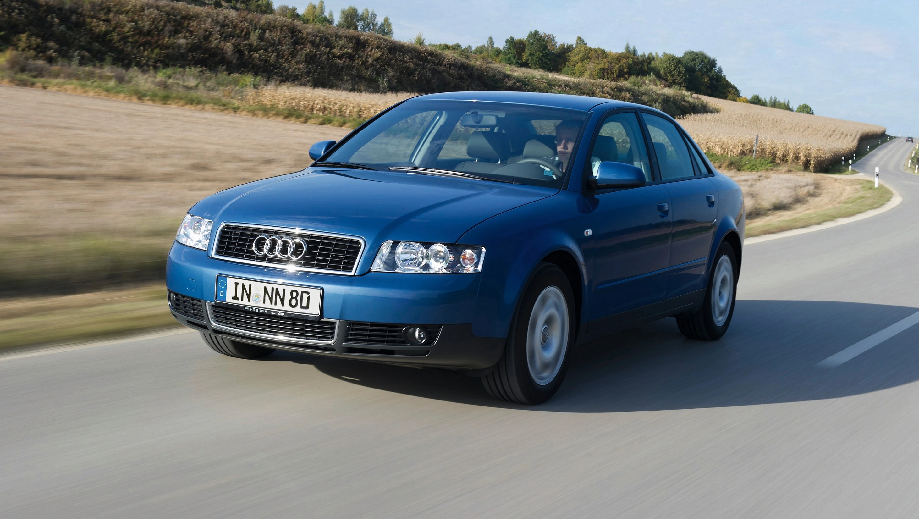 Blauer Audi fährt auf einer Landstraße durch eine ländliche Gegend. Im Hintergrund sind Felder und Bäume zu sehen.