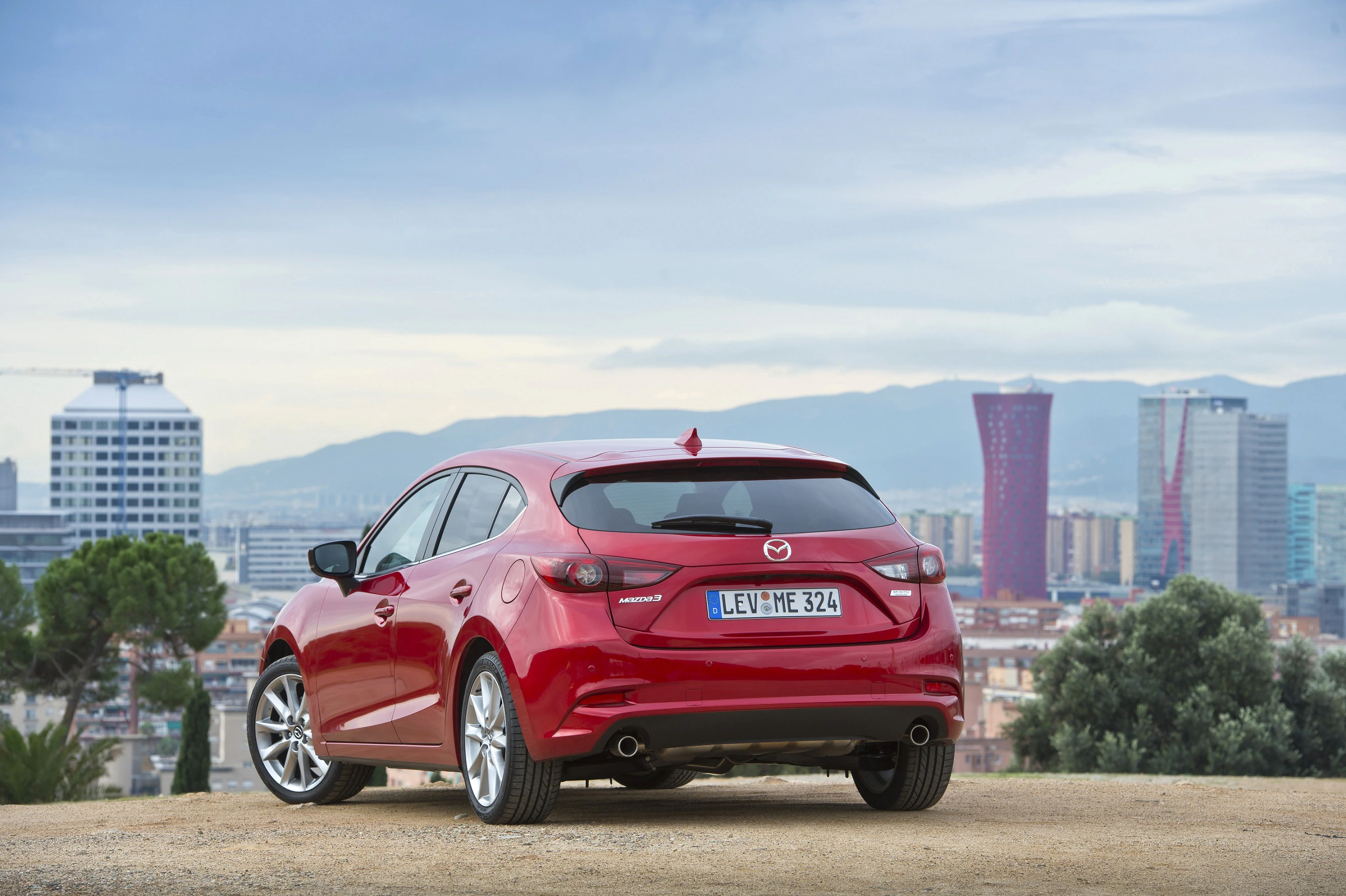Roter Mazda 3 steht auf einem Hügel mit Blick auf eine Stadt mit modernen Gebäuden und Bergen im Hintergrund.