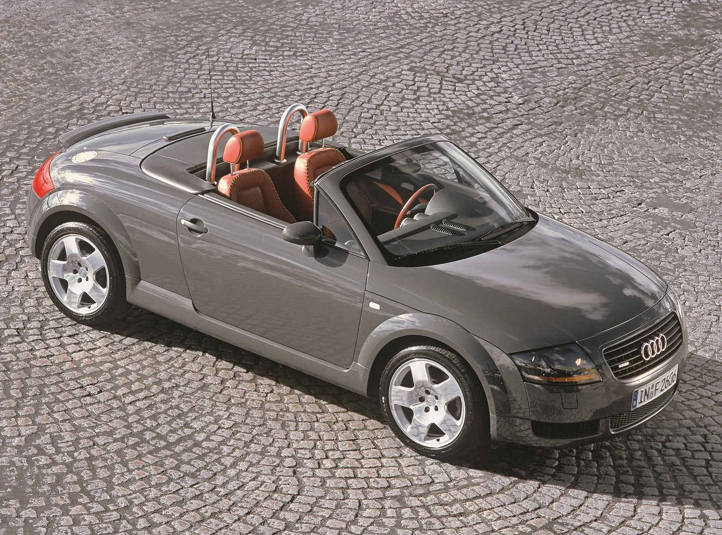 Grauer Audi TT Cabriolet mit roten Ledersitzen, geparkt auf gepflasterter Straße. Draufsicht zeigt elegantes Design und offene Verdeck.
