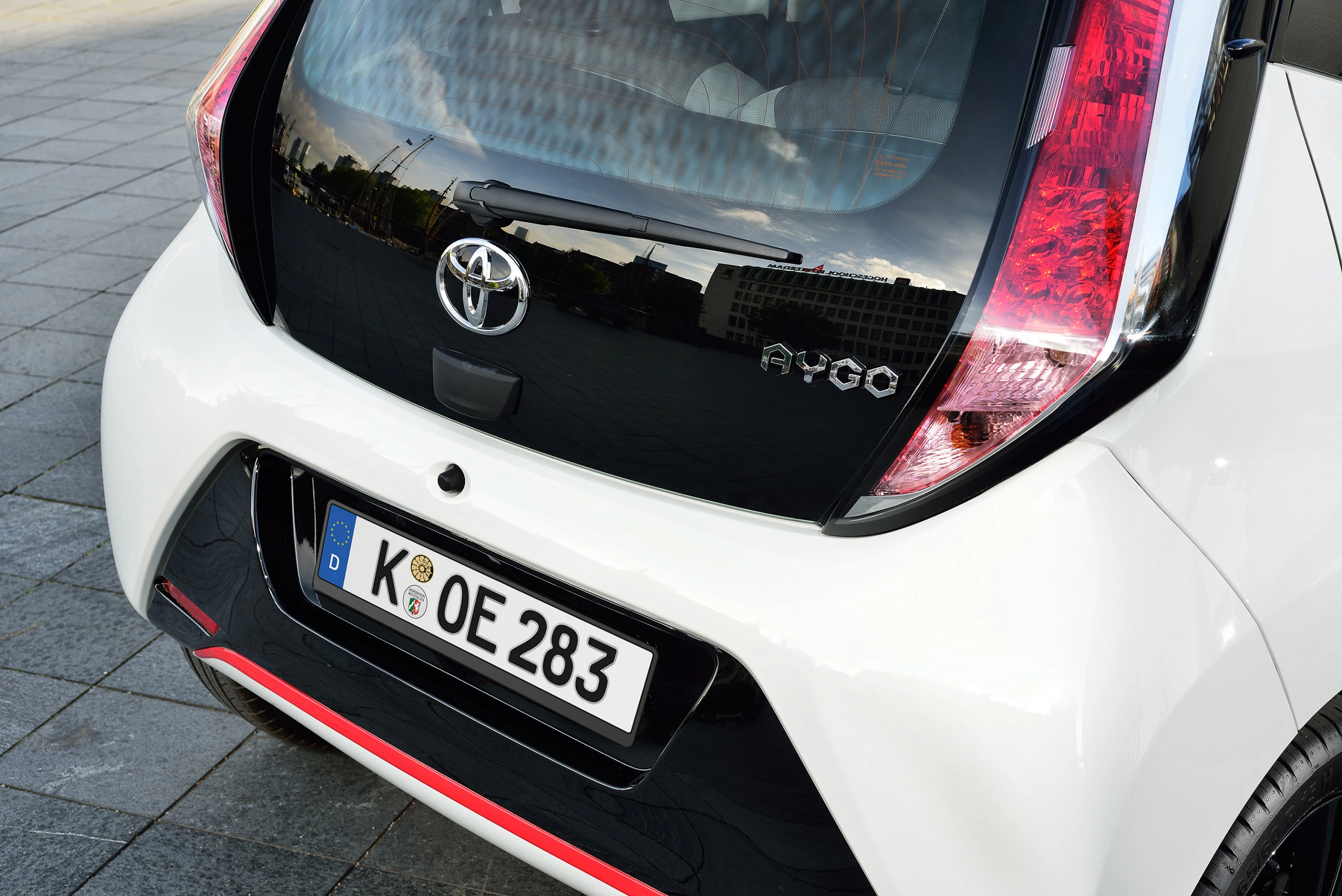 Nahaufnahme eines weißen Toyota Aygo-Hecks mit einem deutschen Nummernschild. Das Auto steht auf einem gepflasterten Platz.