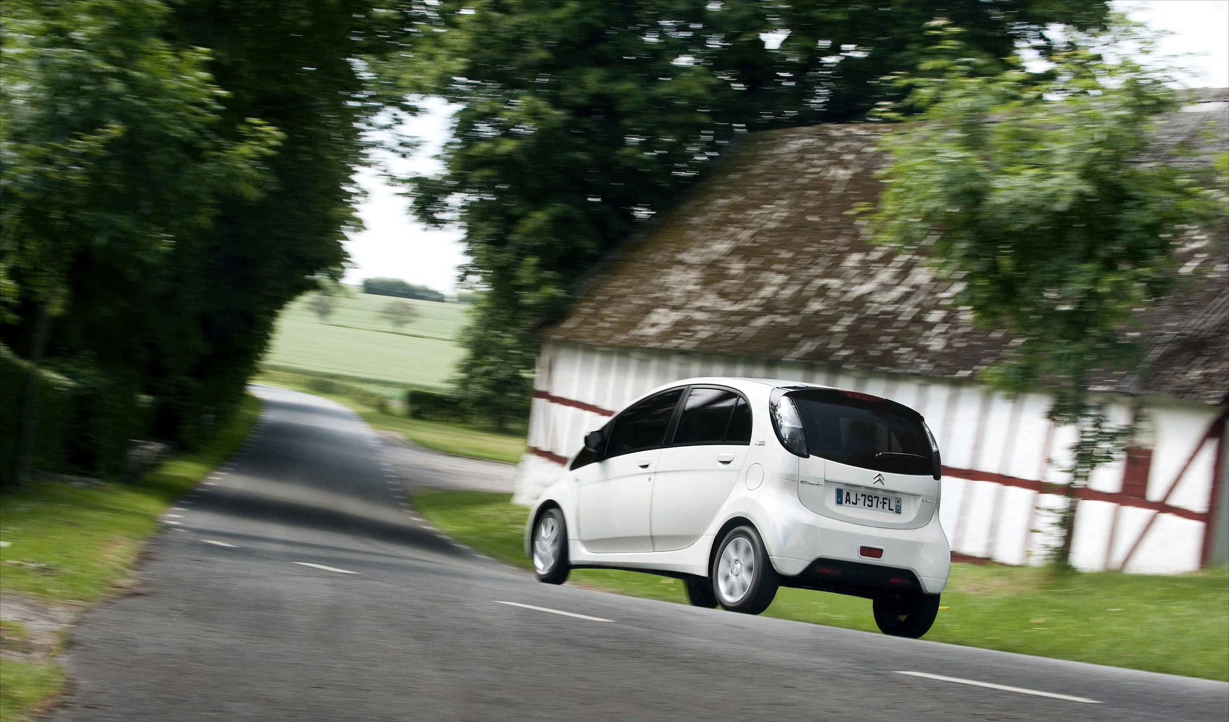 Ein weißes Auto fährt auf einer ländlichen Straße vorbei an einem traditionellen Gebäude mit rotem Dach und weißen Wänden, umgeben von Bäumen.
