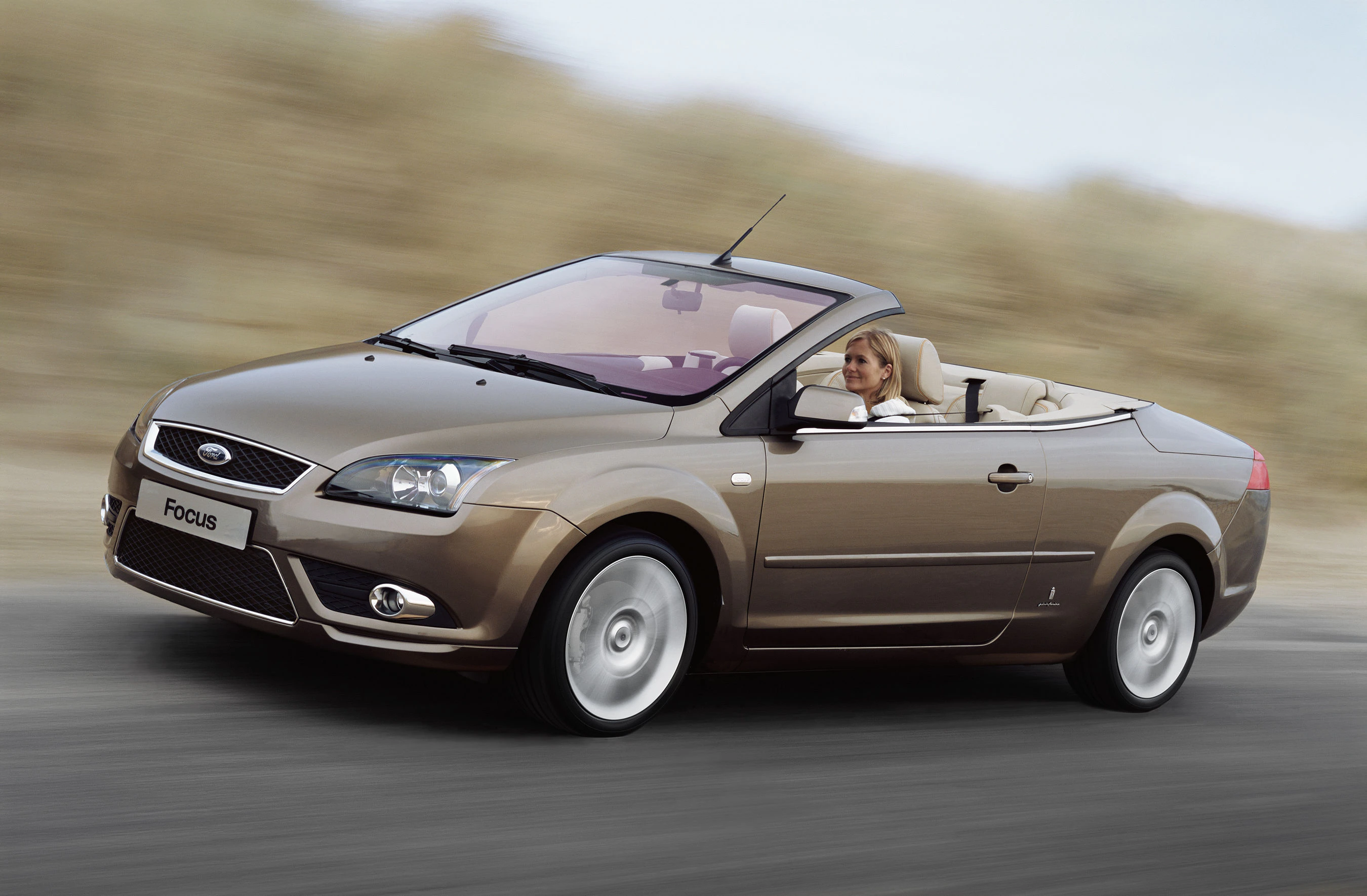 Braunes Ford-Cabriolet in Bewegung, fährt auf einer Straße. Im Auto sitzt eine lächelnde Person. Hintergrund unscharf, sieht aus wie eine sandige Landschaft.