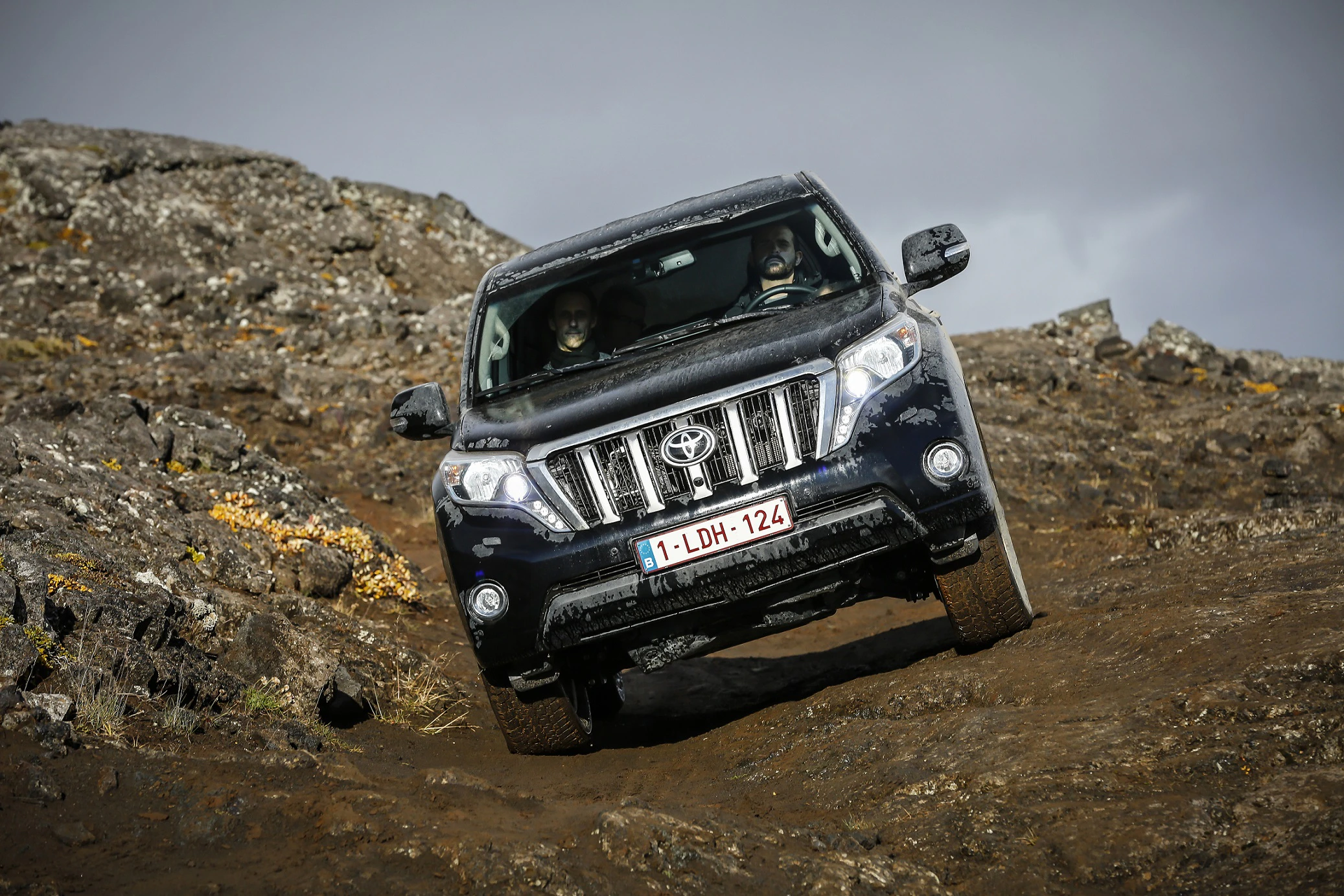 Ein schwarzer SUV fährt auf einer felsigen, steilen Piste hinunter. Der Himmel ist bedeckt, und die Umgebung wirkt rau und unwirtlich.