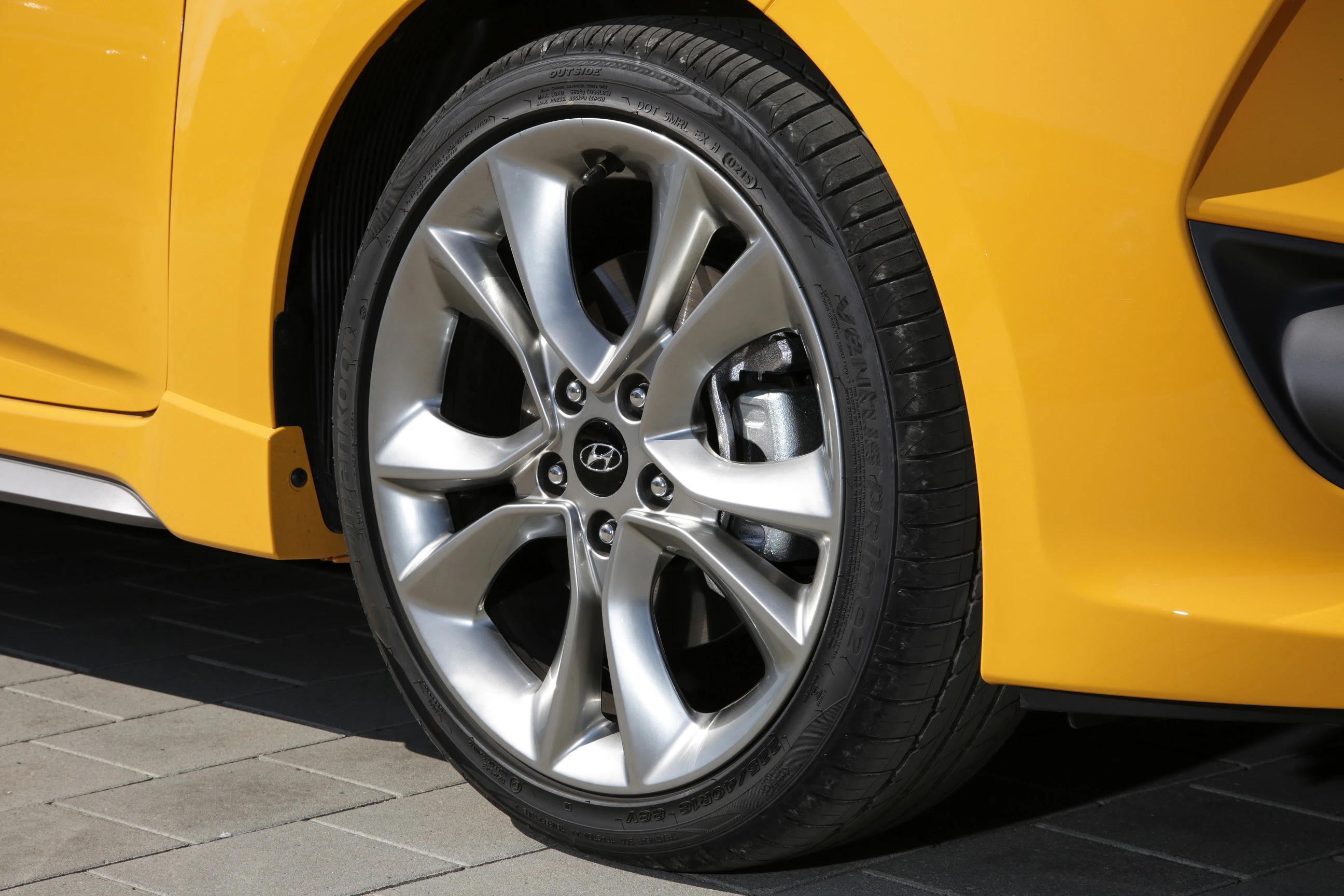 Nahaufnahme eines silbernen Autoreifens mit stilvoller Felge an einem gelben Auto auf gepflastertem Untergrund.