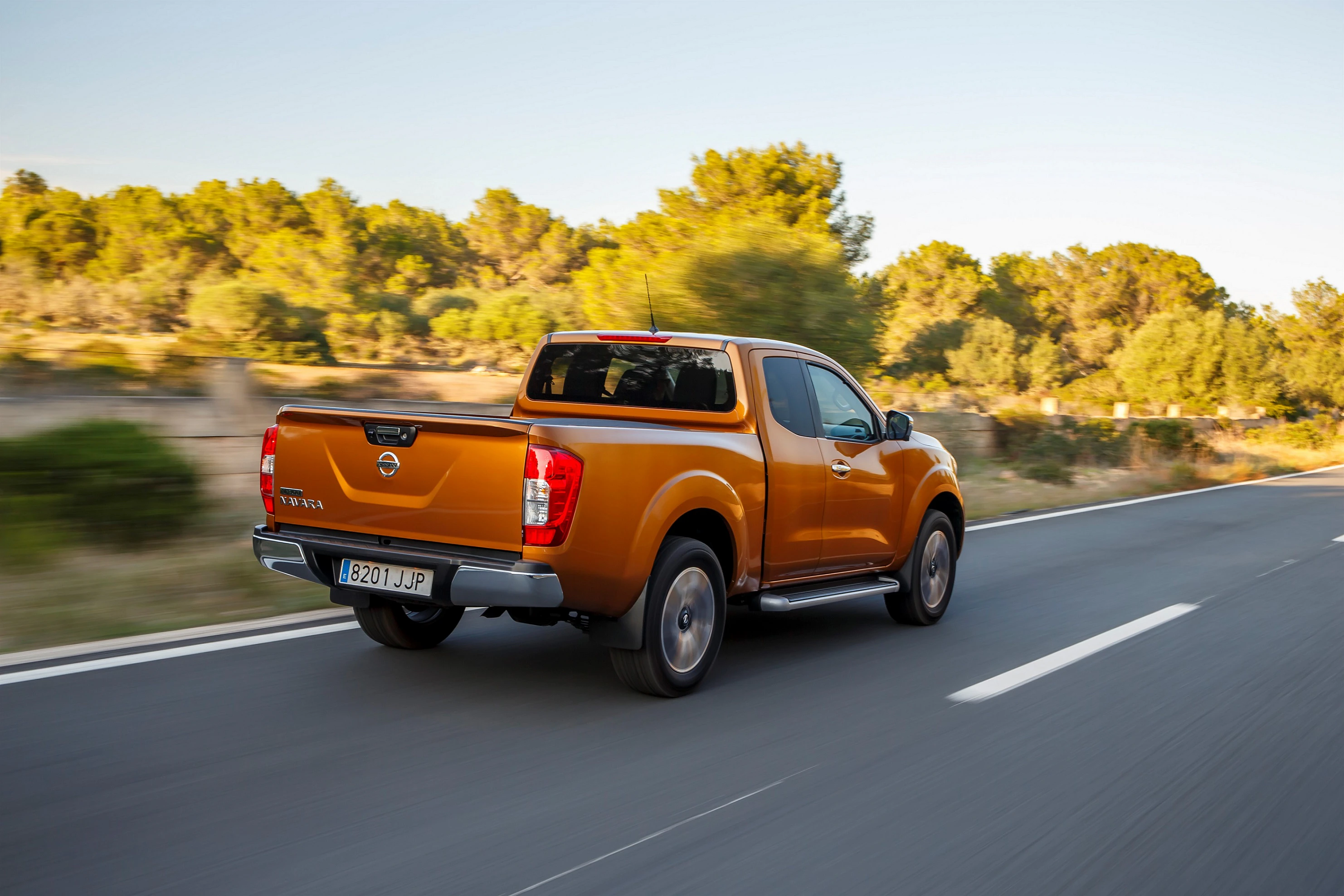 Ein orangefarbener Pickup-Truck fährt auf einer Landstraße, umgeben von Bäumen und blauem Himmel. Nummernschild: 8270 JUP.