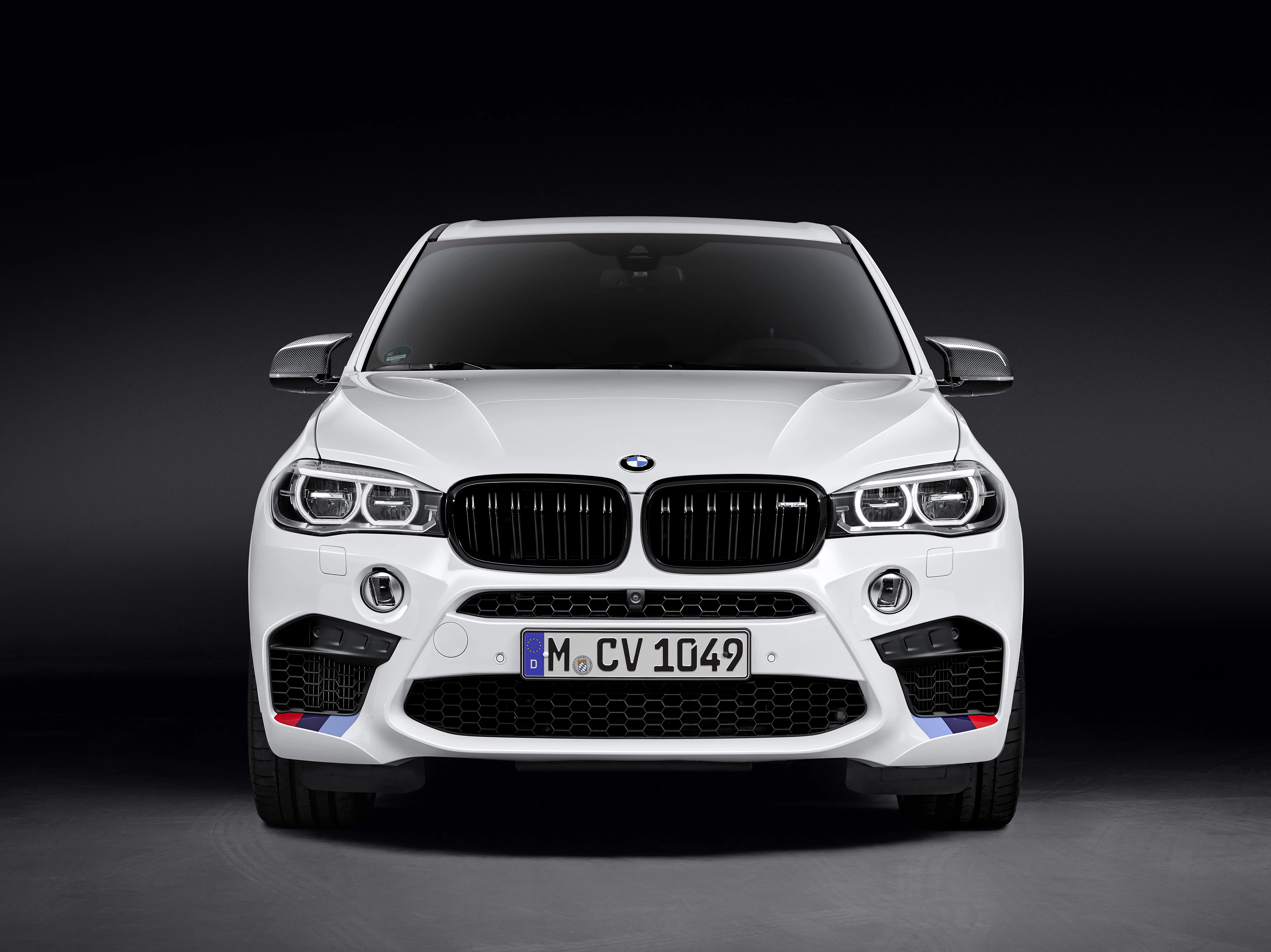 Weißer BMW X5 M von vorne mit auffälligen Scheinwerfern und markantem Kühlergrill, vor schwarzem Hintergrund.