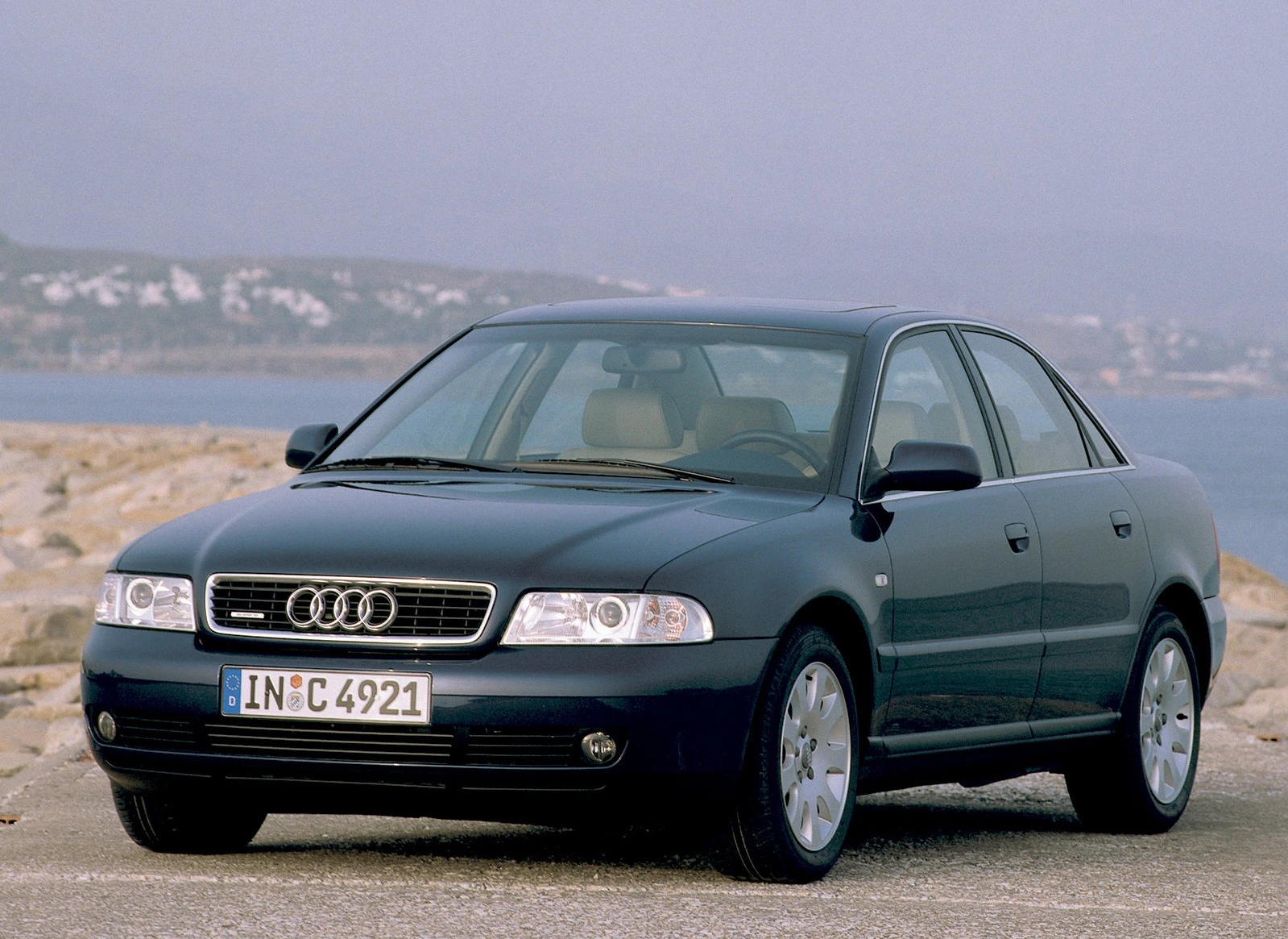 Silberner Audi-Limousine parkt auf einer Küstenstraße, im Hintergrund das Meer und ein bewölkter Himmel.