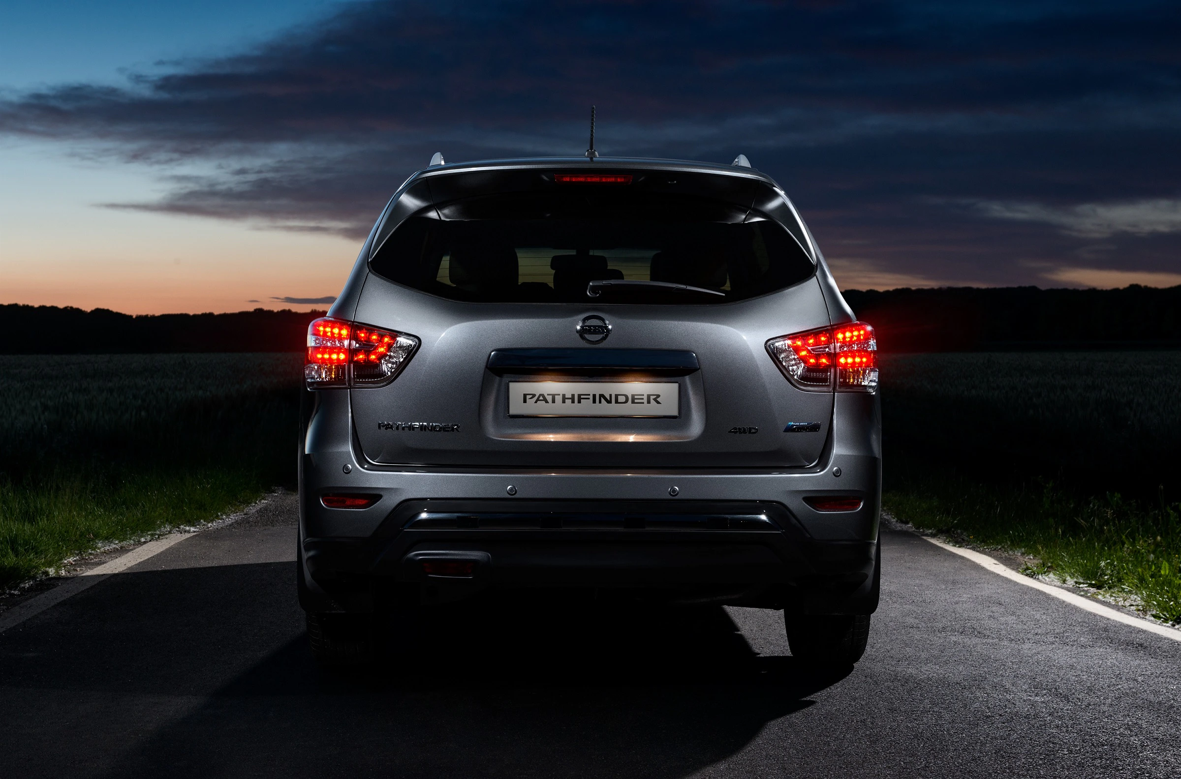 Silberner SUV auf Landstraße, Rückansicht bei Dämmerung mit eingeschalteten Rücklichtern, dunkelblauer Himmel im Hintergrund.