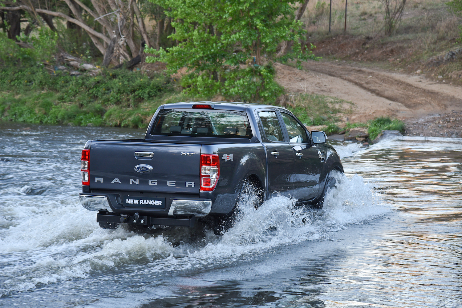 Ein grauer Pickup-Ford Ranger durchquert einen flachen Fluss, spritzendes Wasser, umgeben von Bäumen und Natur.