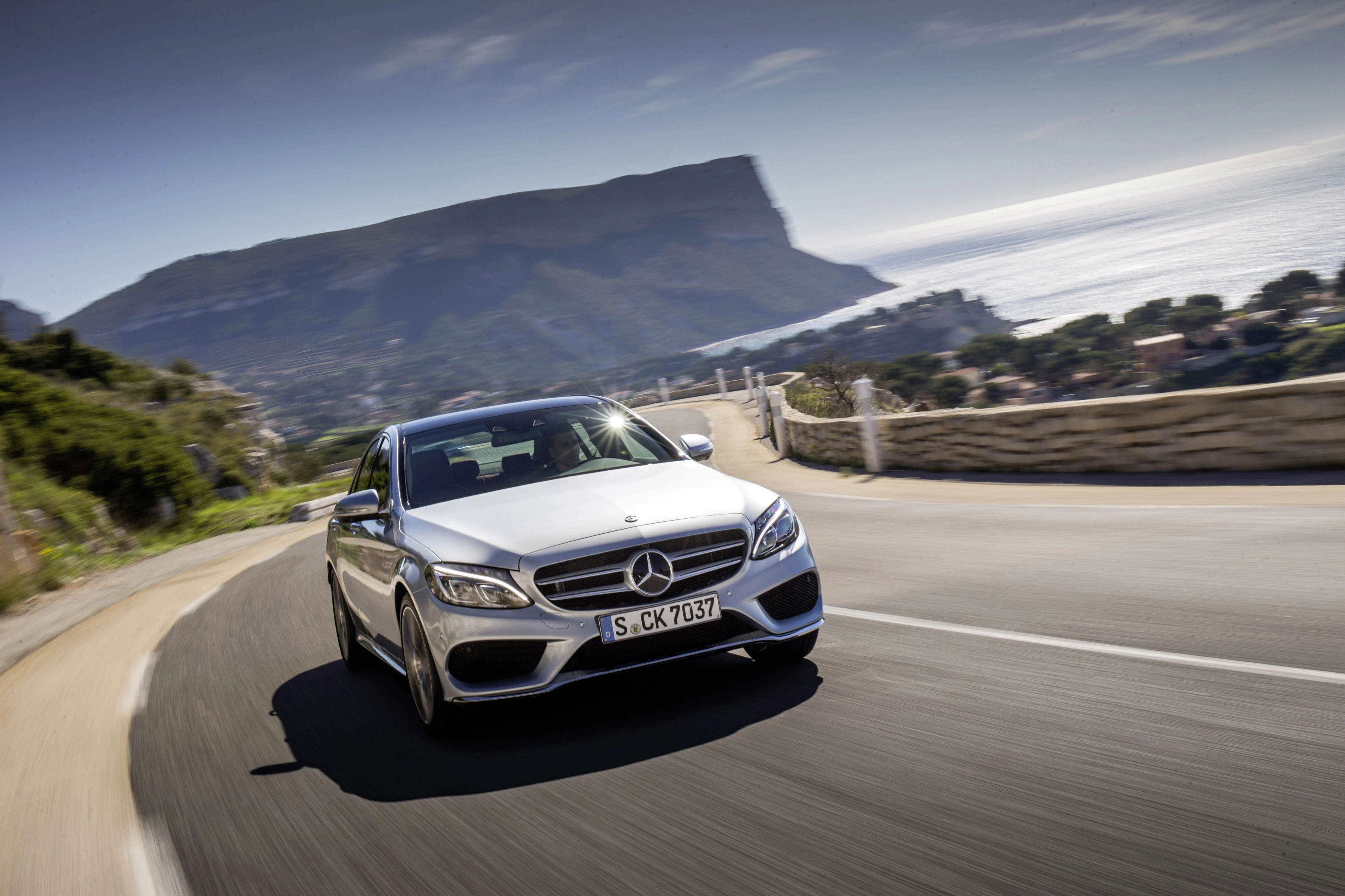 Silberner Mercedes-Benz fährt auf Küstenstraße mit Blick auf Meer und Berge bei sonnigem Wetter.