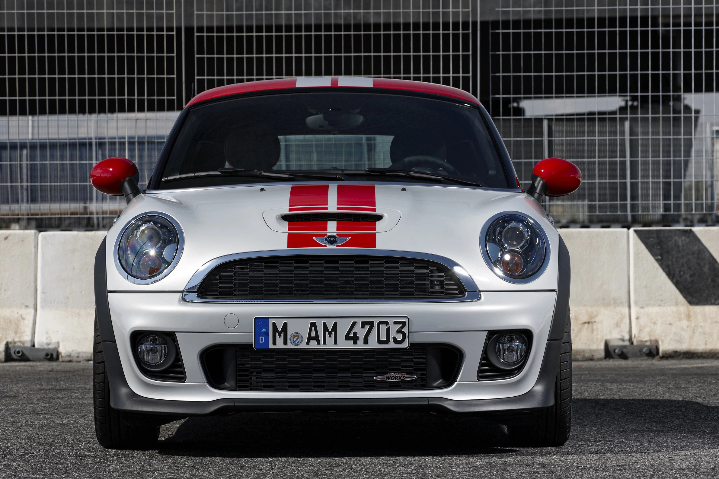 Silberner Mini Cooper mit roten Rennstreifen, Frontansicht vor einer Betonbarriere, leeres Rennstrecken-Umfeld.