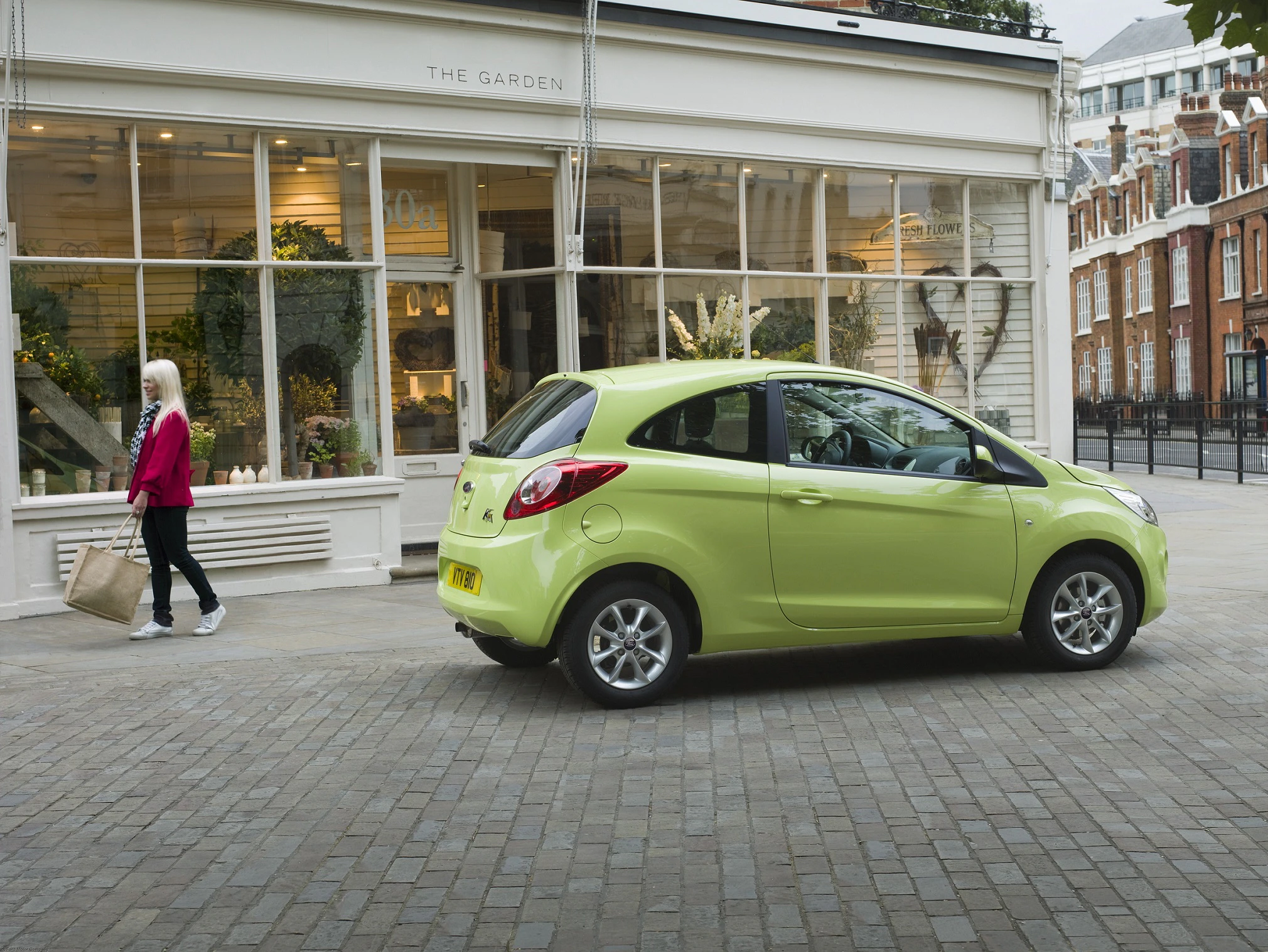 Ein grünes Auto parkt vor einem Blumenladen. Eine Person mit weißen Haaren und einer Einkaufstasche geht vorbei.