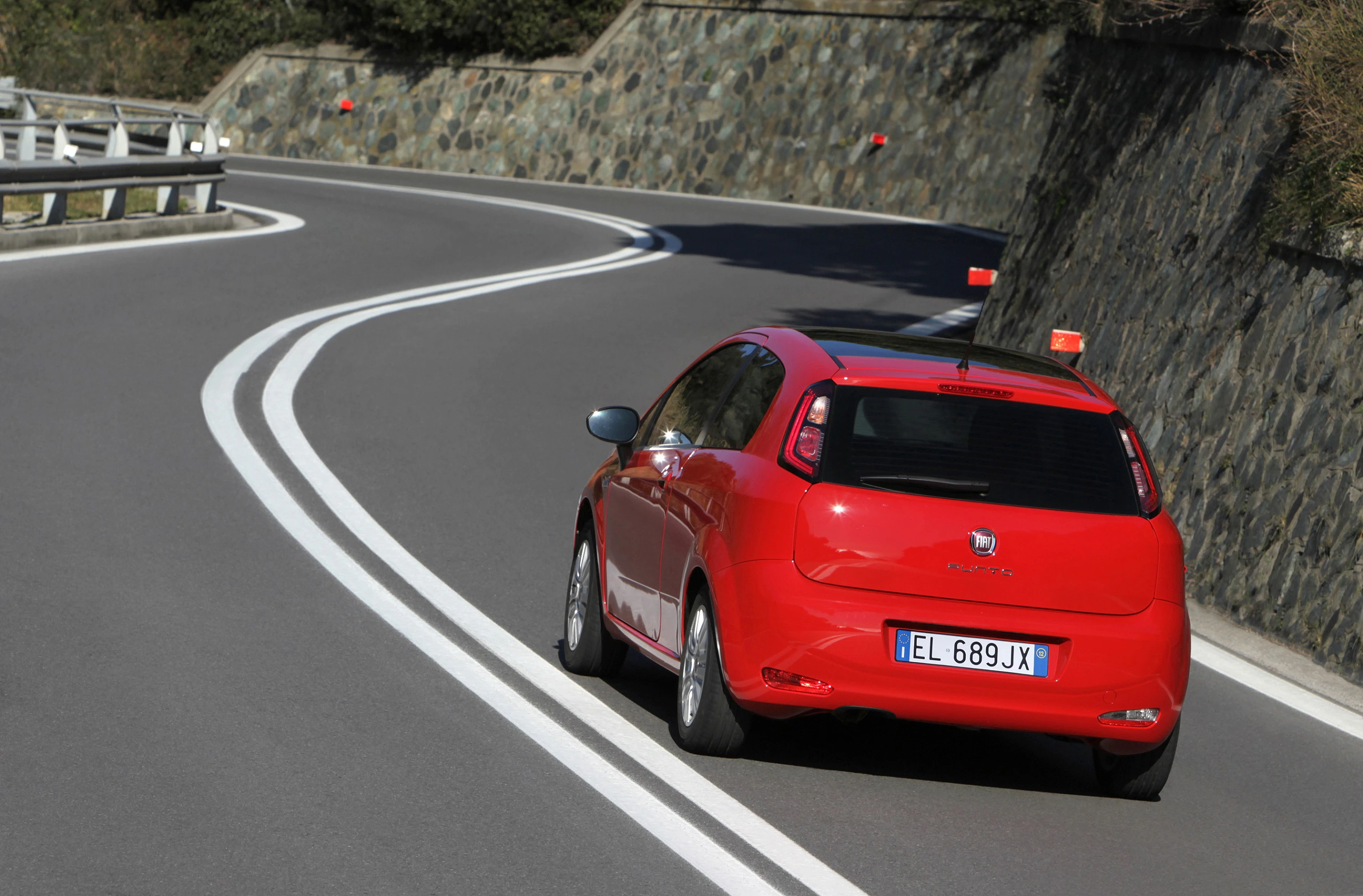 Ein rotes Auto fährt auf einer kurvigen Bergstraße entlang einer Steinmauer. Die Straße hat doppelte weiße Linien.