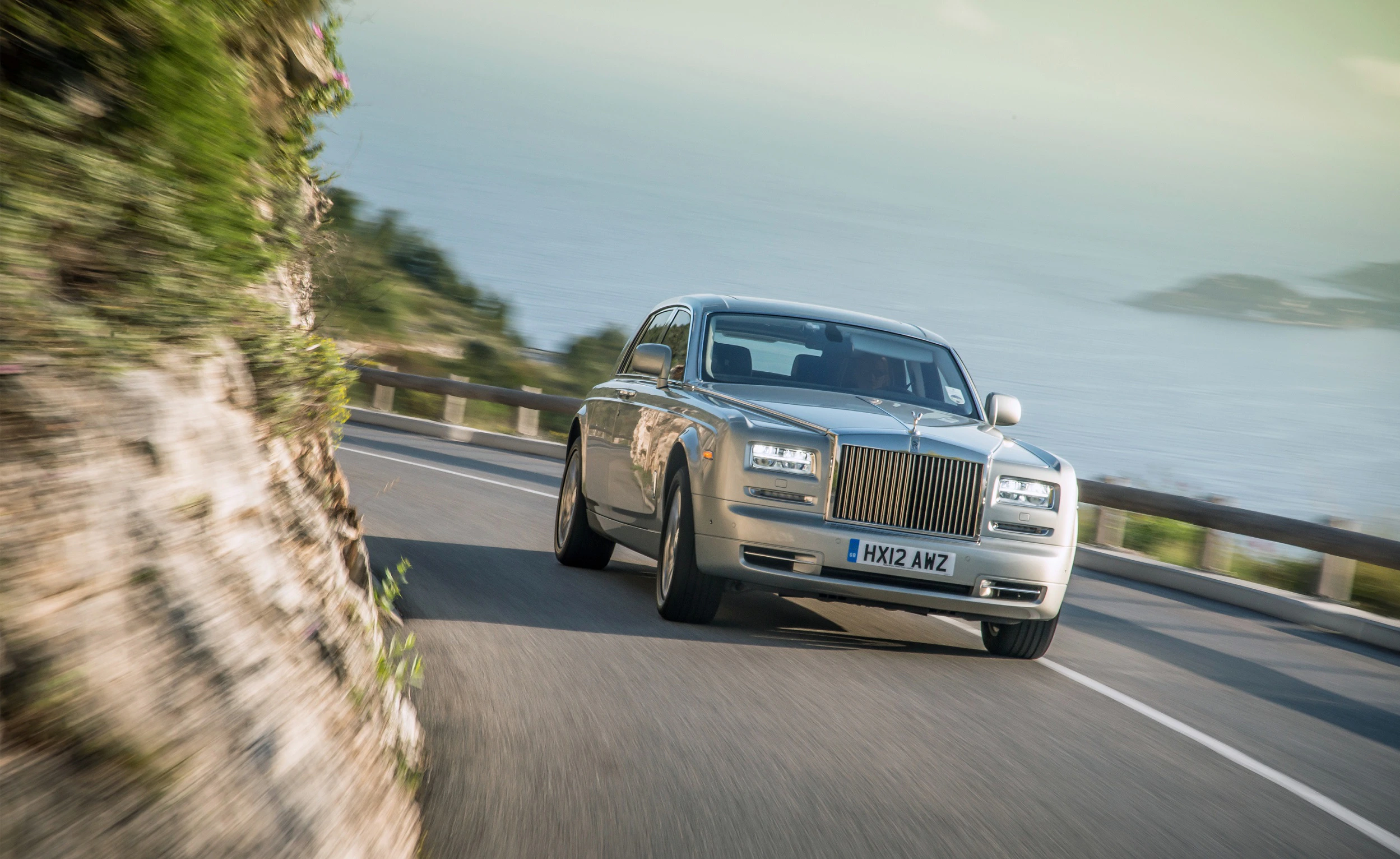 Luxuriöser silberner Rolls-Royce auf kurviger Küstenstraße mit Meerblick im Hintergrund.