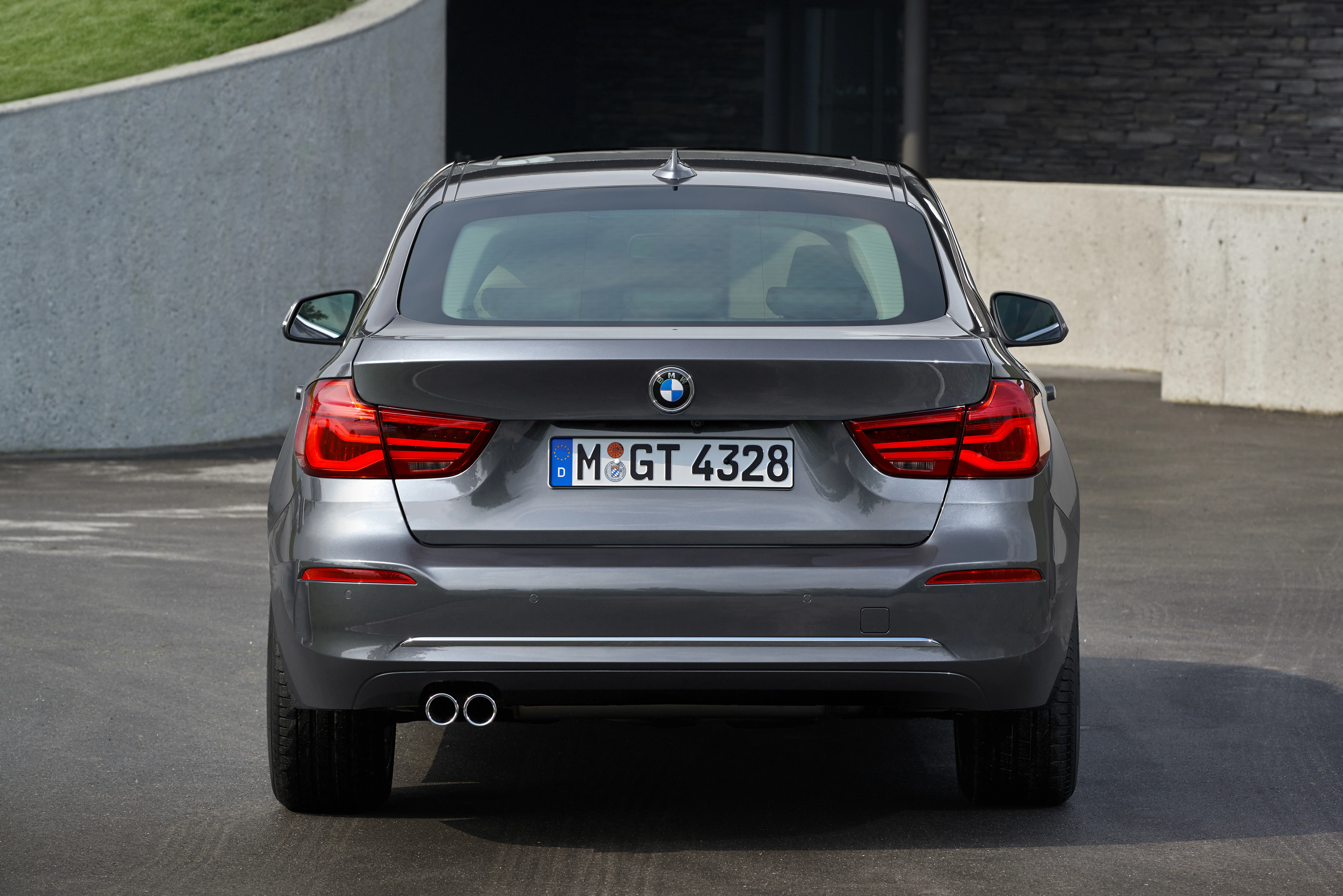 Rückansicht eines grauen BMW auf einer Straße, vor einer Betonmauer geparkt. Fahrzeug hat roten Rückleuchten und zwei Auspuffrohre.