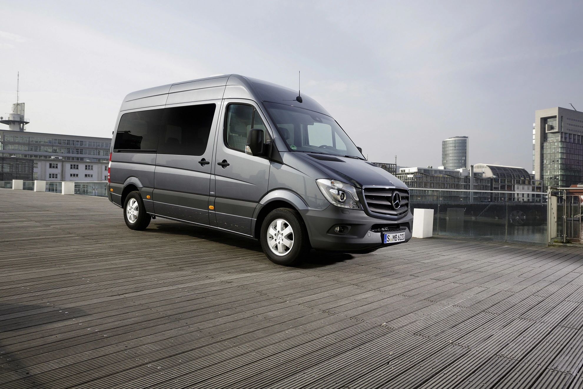 Grauer Mercedes-Benz Sprinter parkt auf Holzplanken vor moderner Architektur und Wasser, hell bewölkter Himmel im Hintergrund.