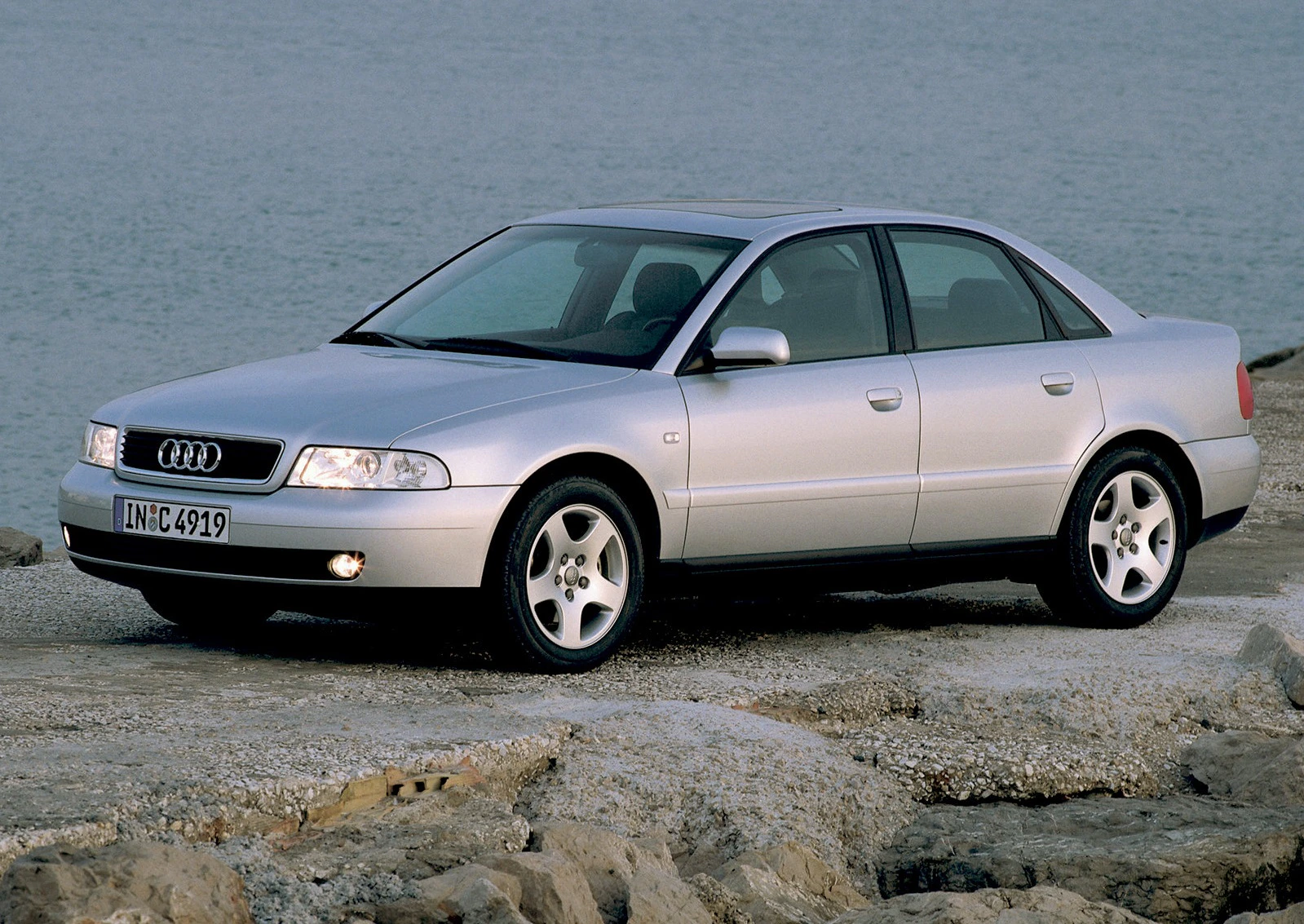 Silberner Audi A4 Limousine auf felsigem Untergrund am Wasser stehend, seitlich fotografiert, mit sanftem Sonnenlicht.