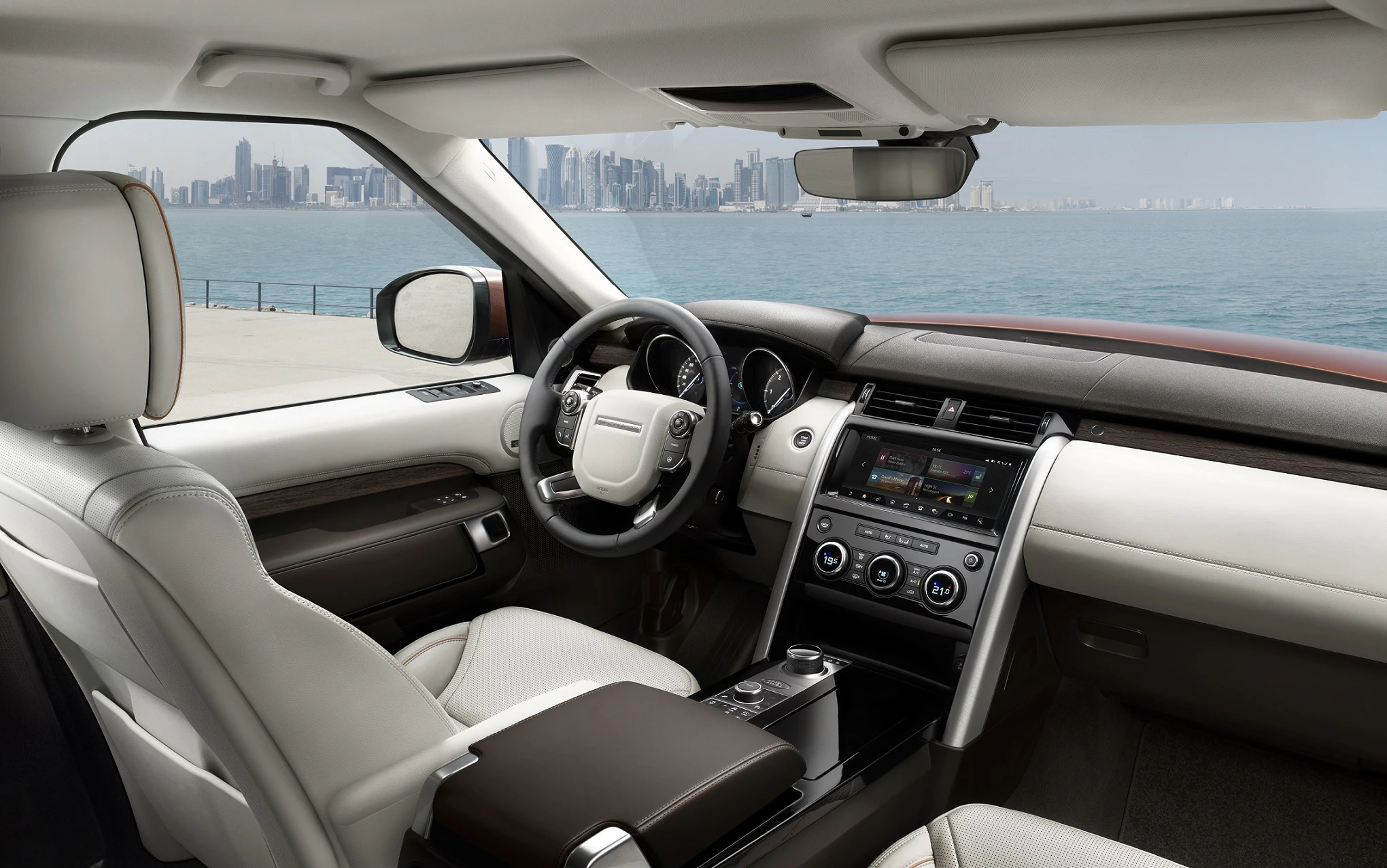 Innenraum eines luxuriösen Autos mit Blick auf das Meer und die Skyline einer Stadt im Hintergrund. Beige Ledersitze und modernes Armaturenbrett.