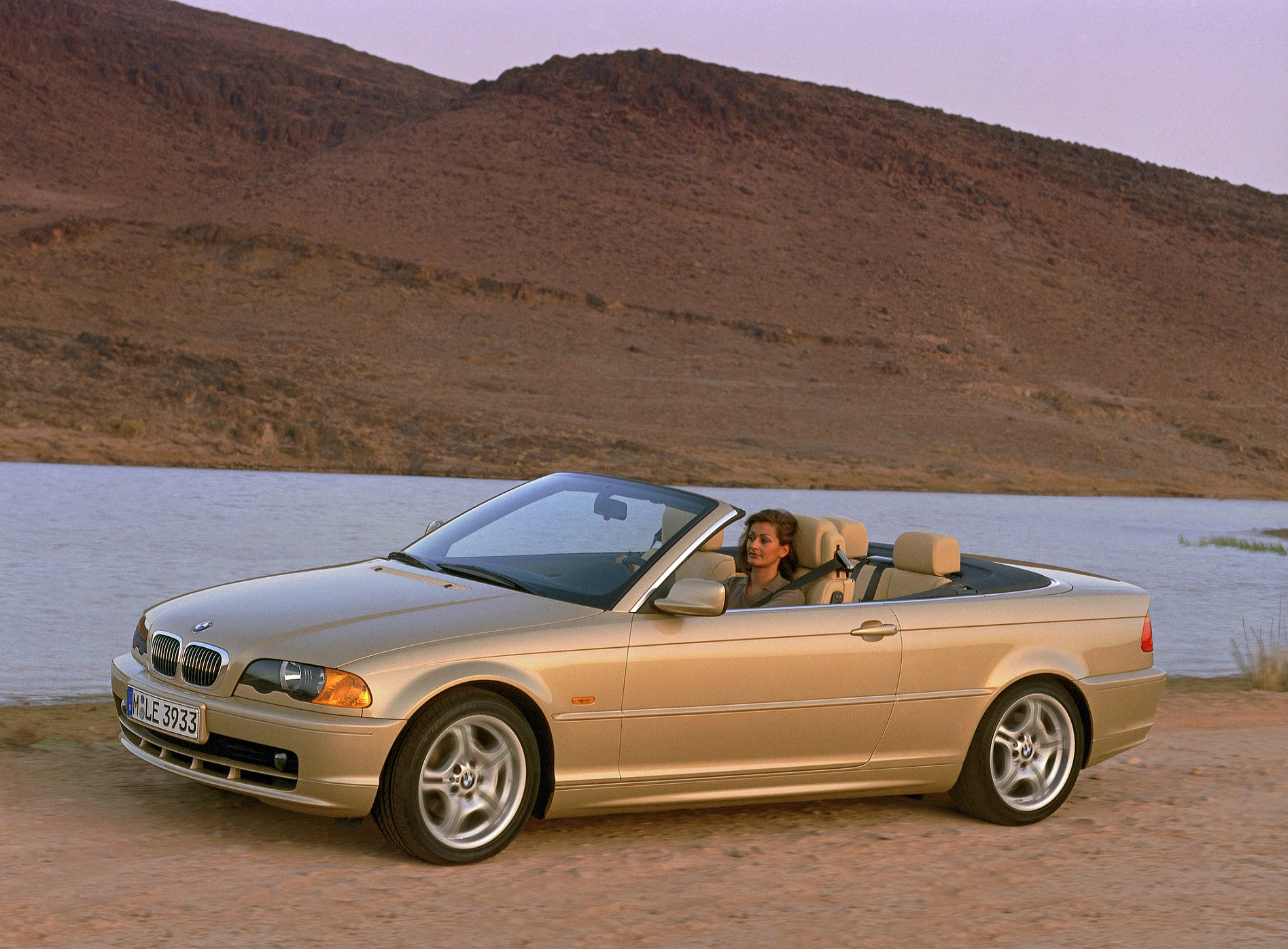 Goldener BMW Cabrio, stehend auf einer sandigen Straße. Im Hintergrund ein See und bergige Landschaft. Eine Person sitzt am Steuer.