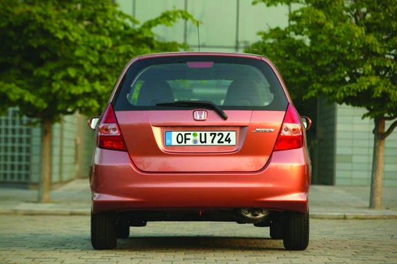 Roter Honda Jazz parkt auf gepflastertem Gelände, umgeben von Bäumen, Gebäude im Hintergrund.
