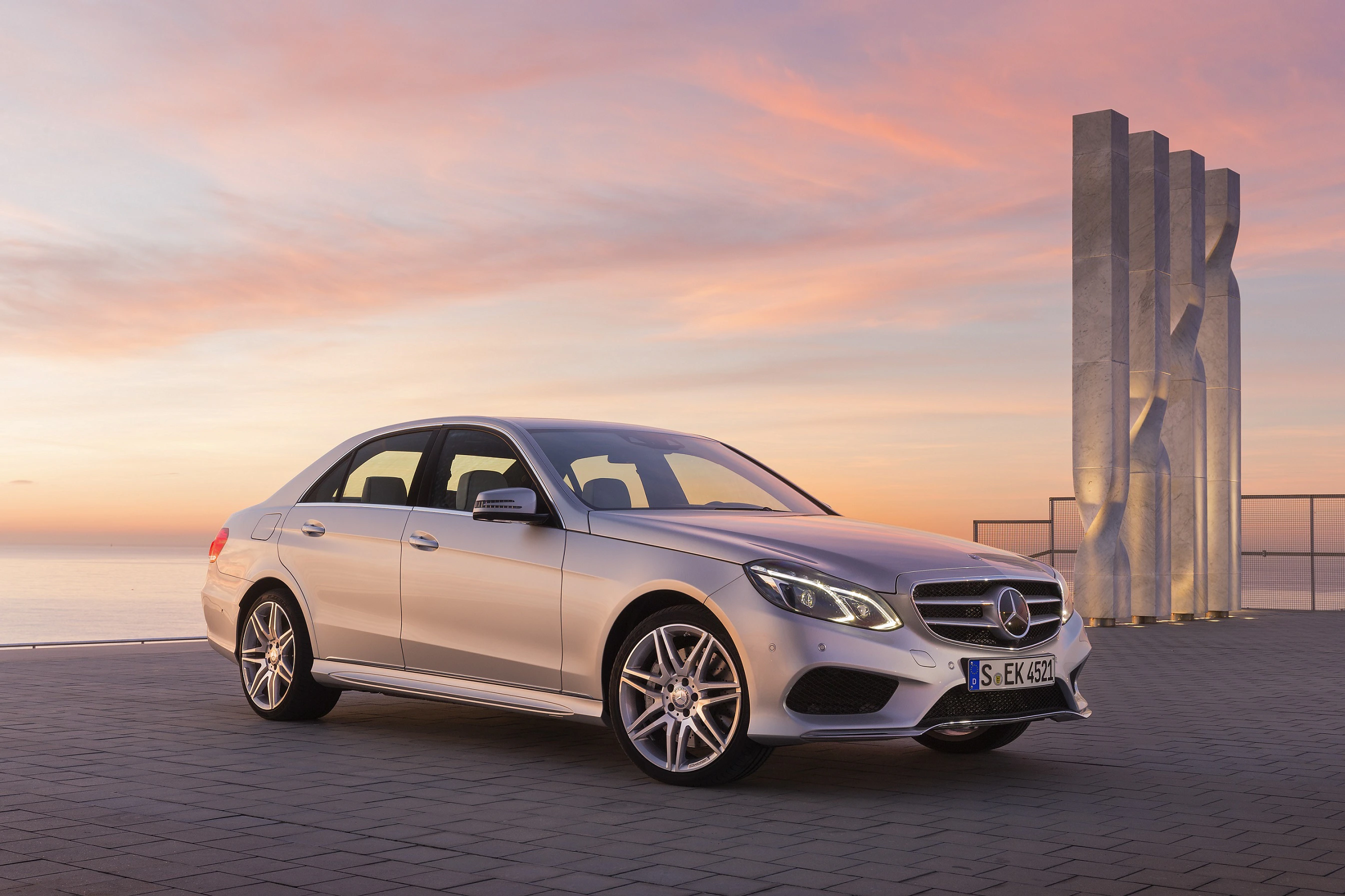 Silberner Mercedes-Benz auf einer Promenade bei Sonnenuntergang, im Hintergrund modernes Kunstwerk und sanfter Himmel in Orange- und Rosatönen.